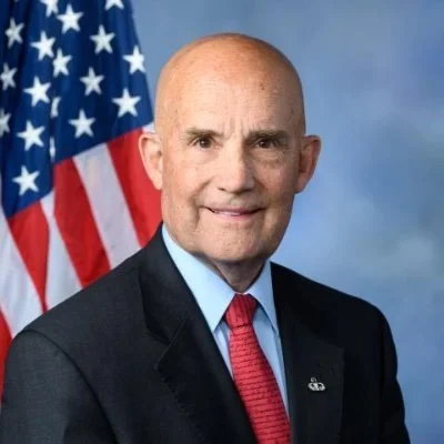 Bald man in a suit standing in front of an American flag.