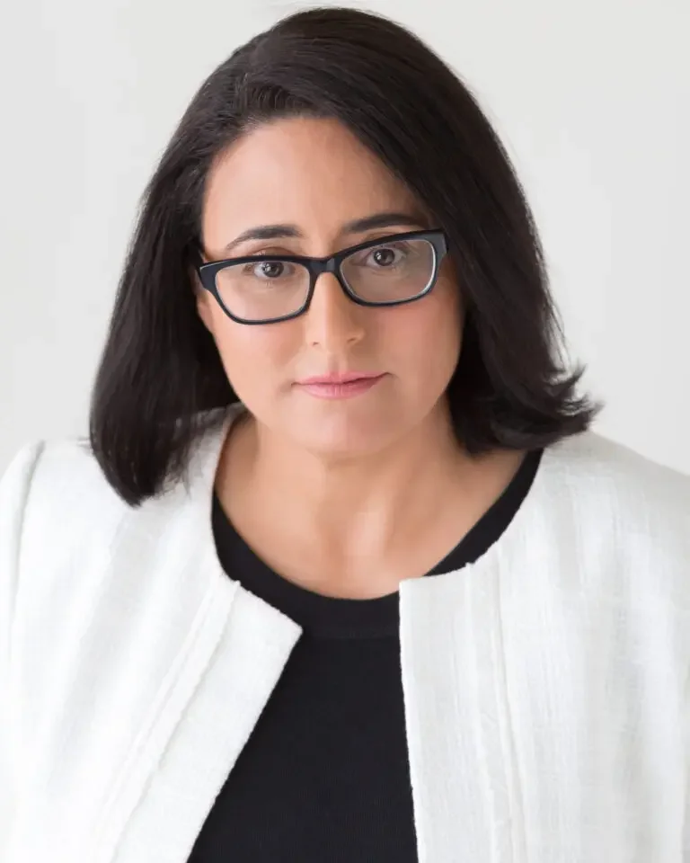 A woman with shoulder-length dark hair and glasses, wearing a black shirt and a white blazer, looking at the camera against a plain light background.