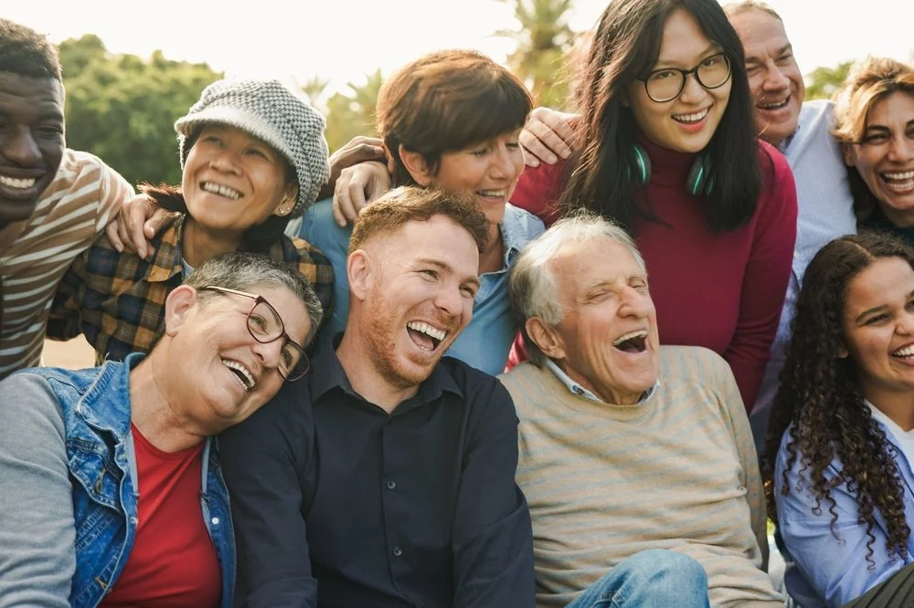 A diverse group of eleven people smiling and laughing outdoors on a sunny day, showing camaraderie and joy.