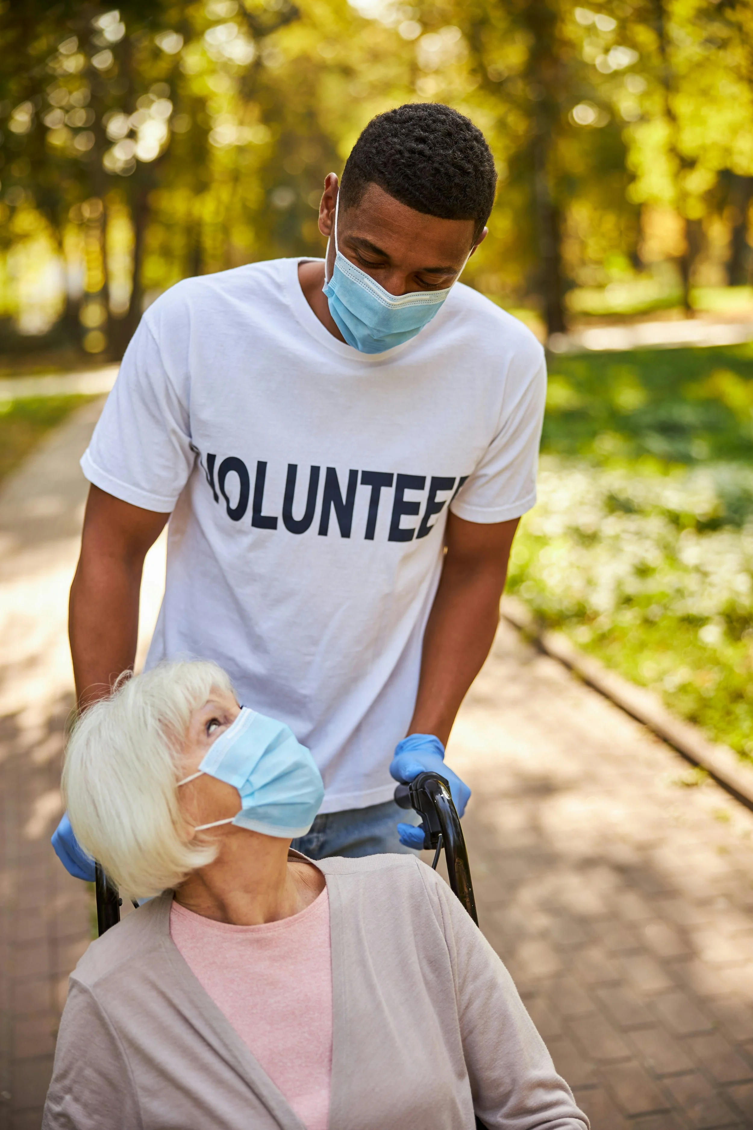A young man wearing a face mask and a volunteer t-shirt pushes an elderly woman in a wheelchair, both wearing face masks, on a pathway in a park with autumn trees.
