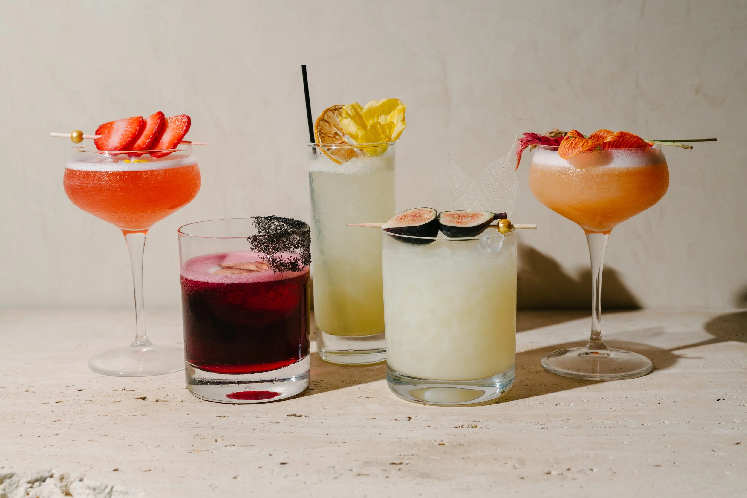 Five colorful cocktails on a light wooden surface, garnished with fruits and decorative elements.