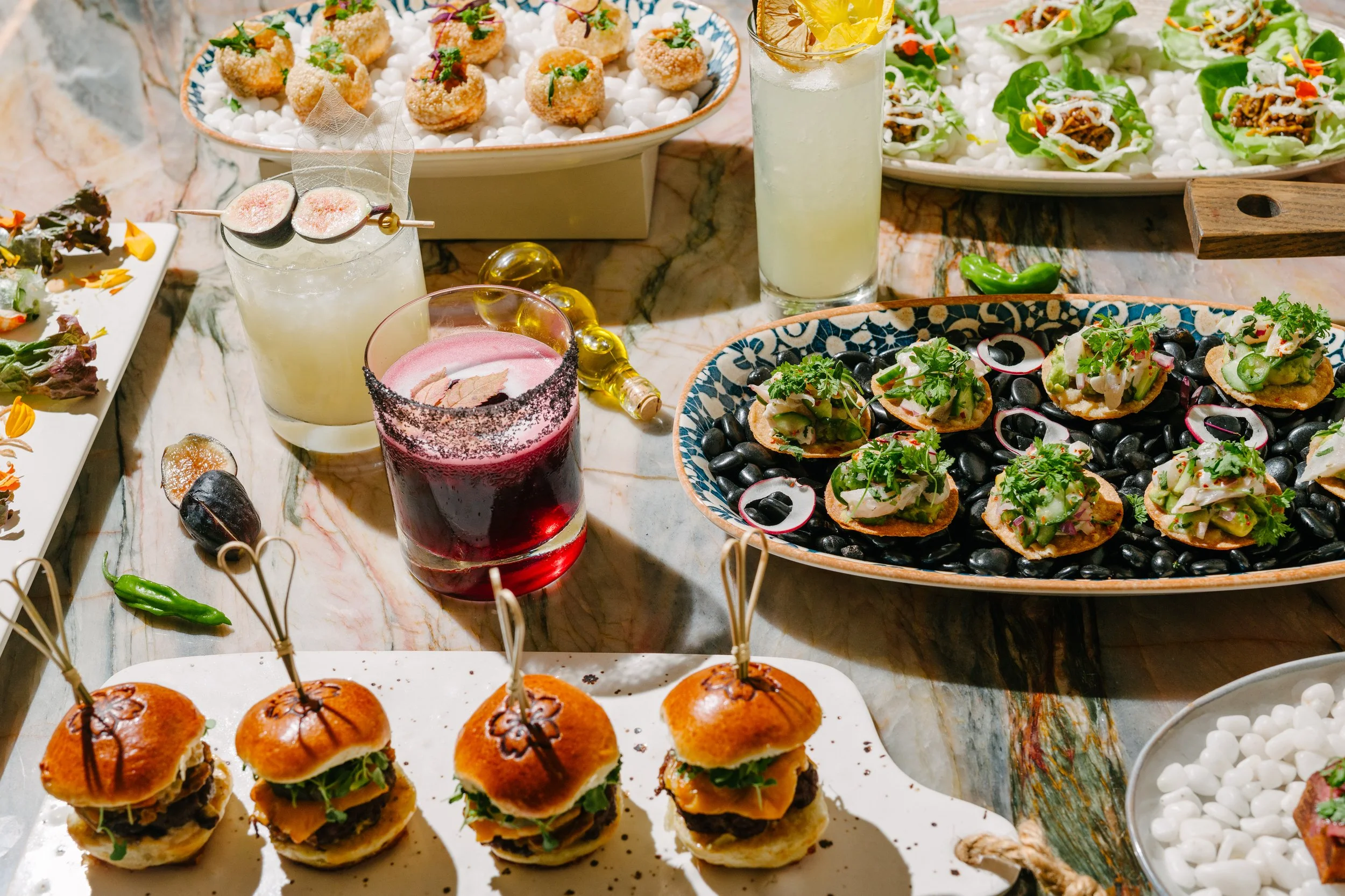 A table with various appetizers and drinks including sliders, small open-faced sandwiches with greens and radishes, and three beverages with colorful garnishes.