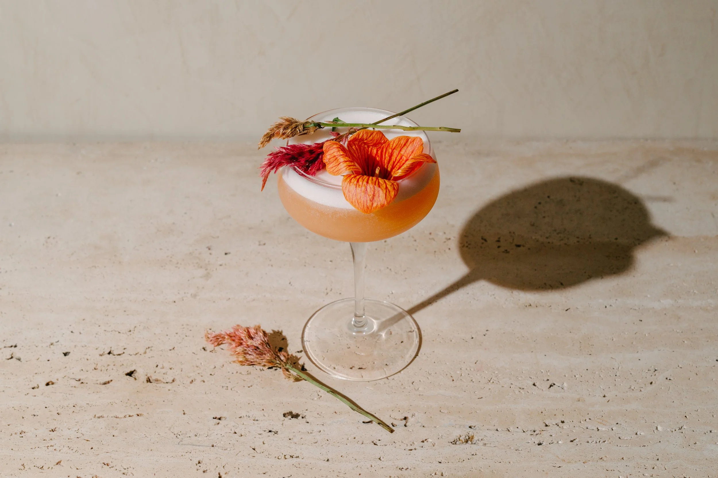 A cocktail in a coupe glass garnished with orange and red flowers and dried grasses, casting a shadow on a light-colored surface.
