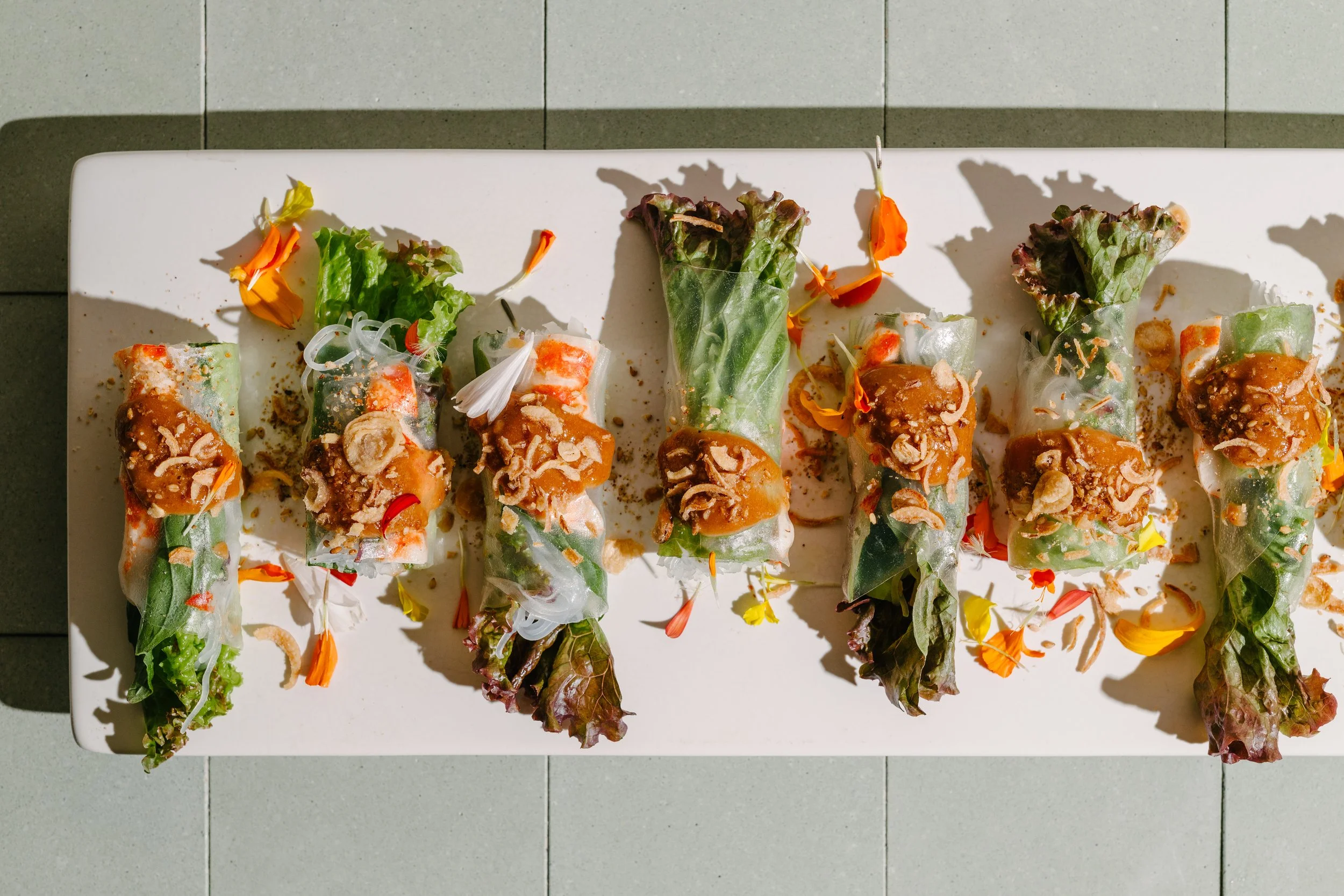 Six spring rolls garnished with sauce and edible flower petals, arranged on a rectangular white platter, with shadows cast on a tiled surface.