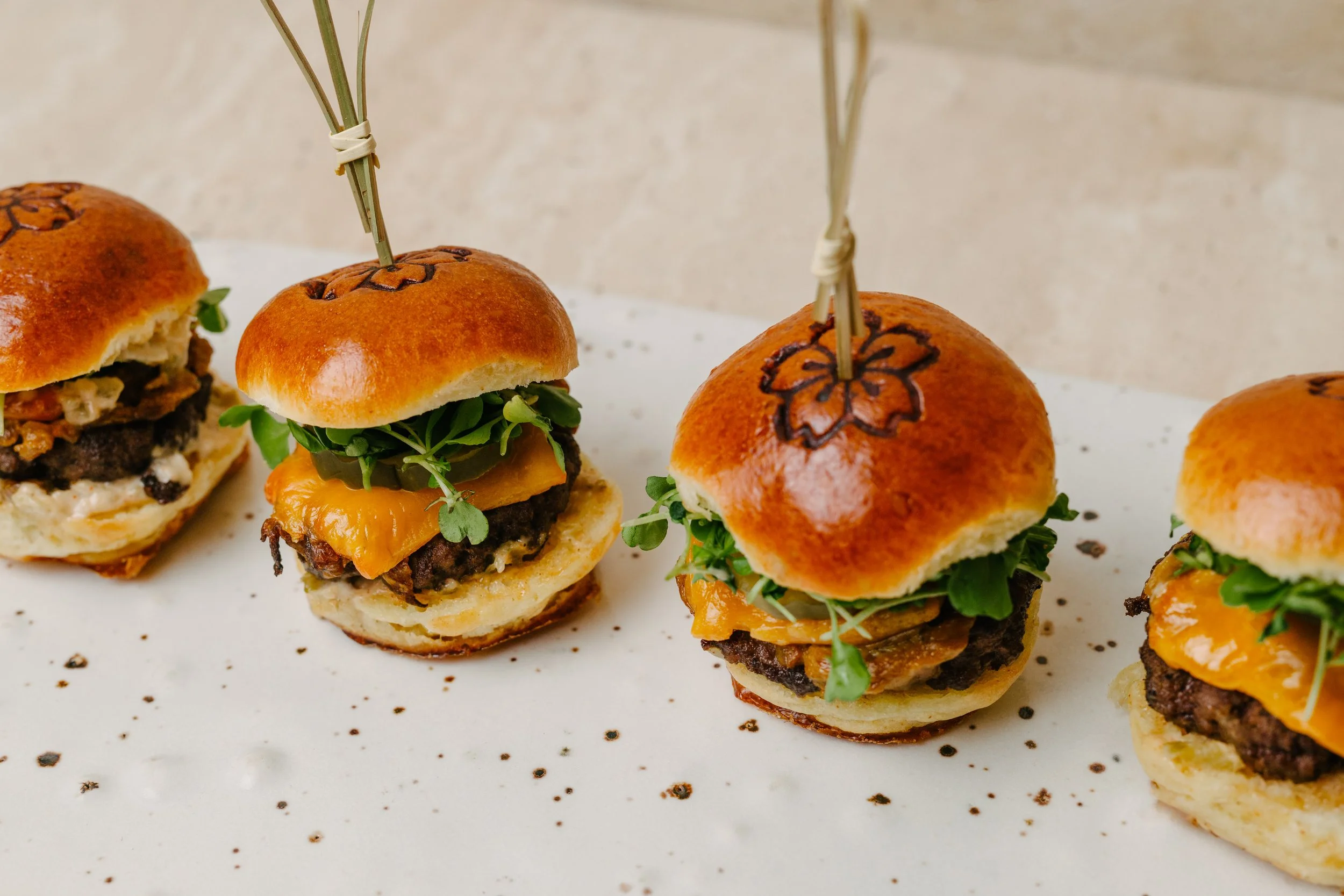 Mini sliders with beef patties, cheddar cheese, green microgreens, and toasted buns, some with decorative grill marks, served on a white platter.