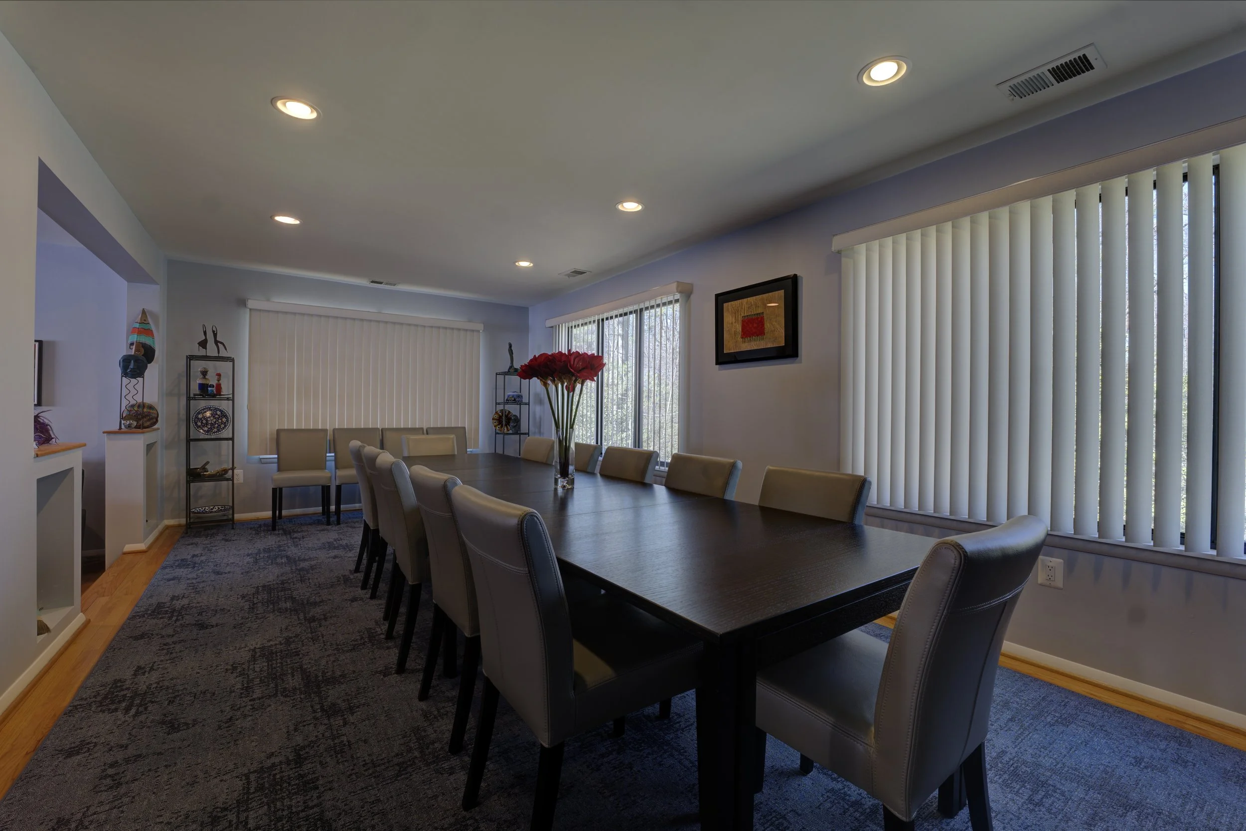 Dining room with a long dark wood table, beige leather chairs, a vase with red flowers, large windows with vertical blinds, and decorated shelves with artwork and sculptures.