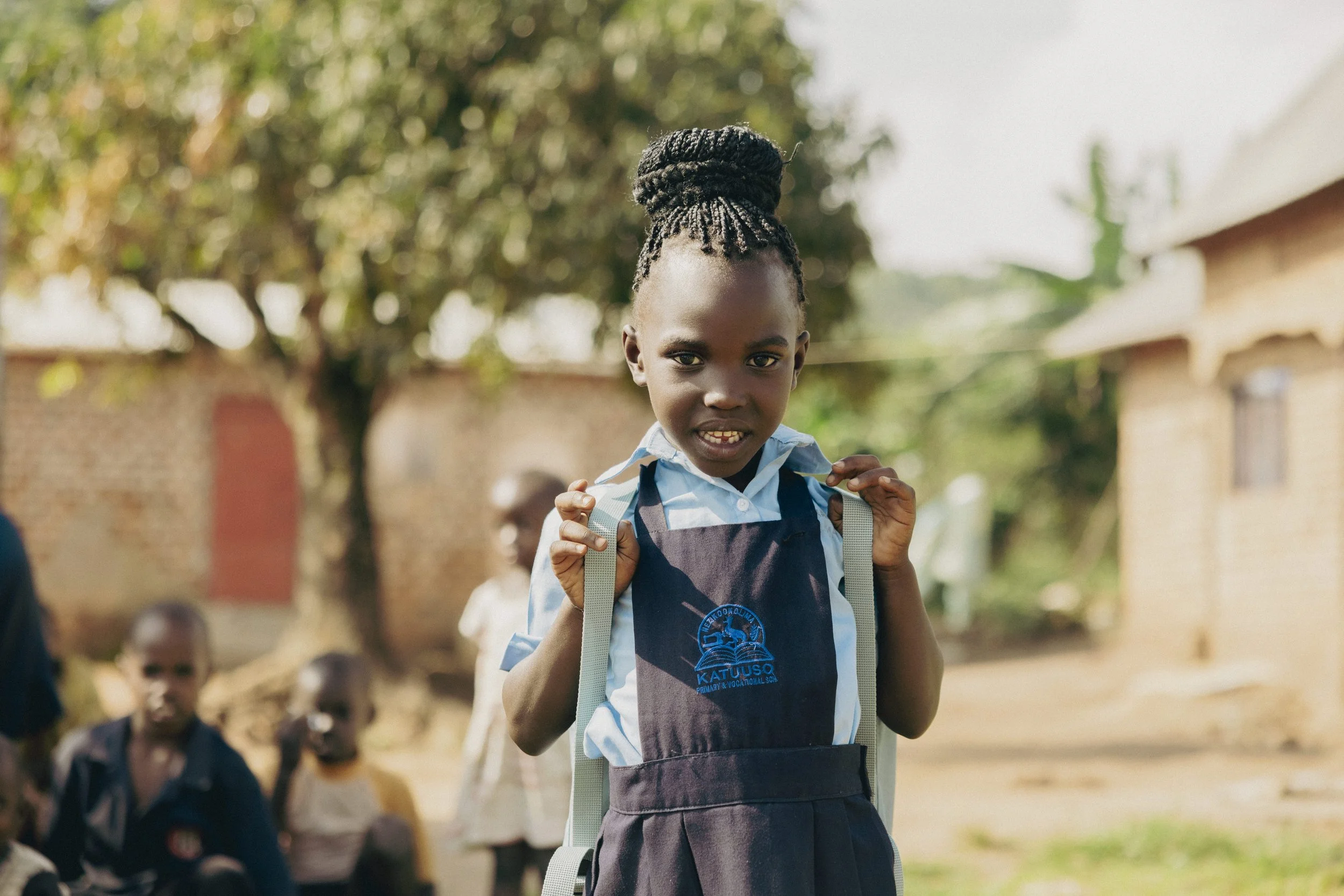 A young girl in school uniform standing outdoors with a backpack on her shoulders, looking at the camera. In the background, other children and a brick building are visible.