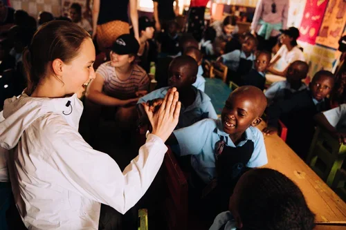 A woman and a group of children in a classroom. The woman is smiling and giving a high-five to a young boy who is also smiling. Other children are seated at desks, engaged and smiling, with colorful posters on the walls.