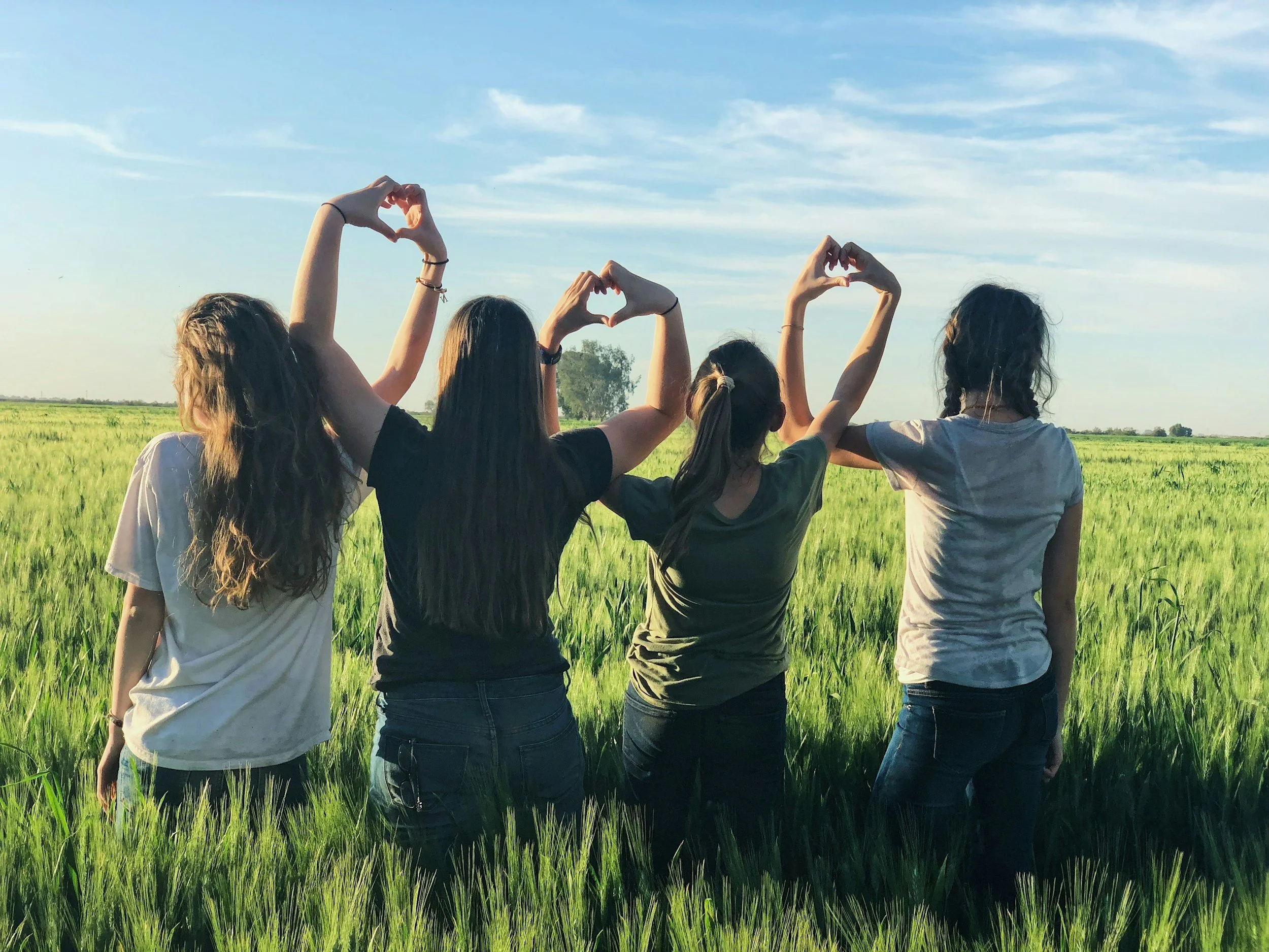 Four women standing in a green field making heart shapes with their hands, facing away from the camera, under a clear blue sky.
