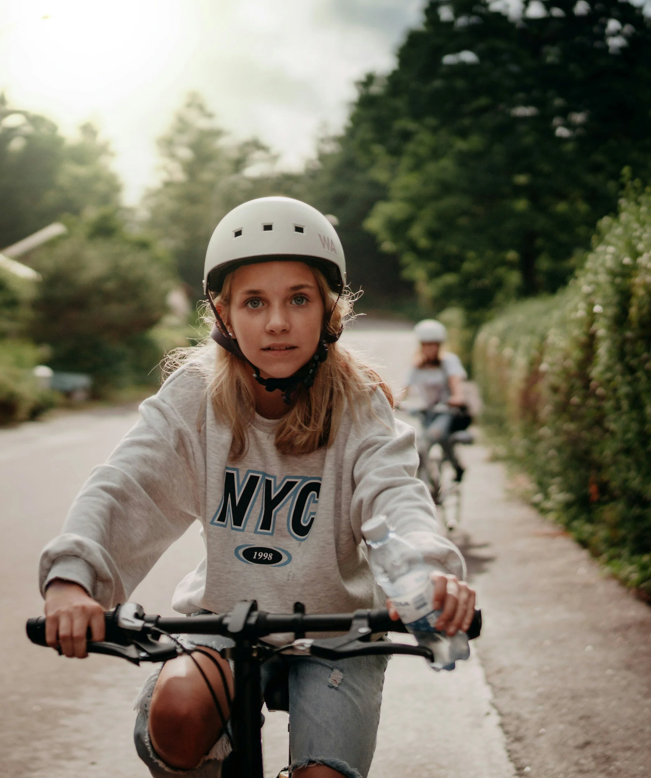 A teen girl in a white NYC sweatshirt is riding a bike and wearing a white helmet.