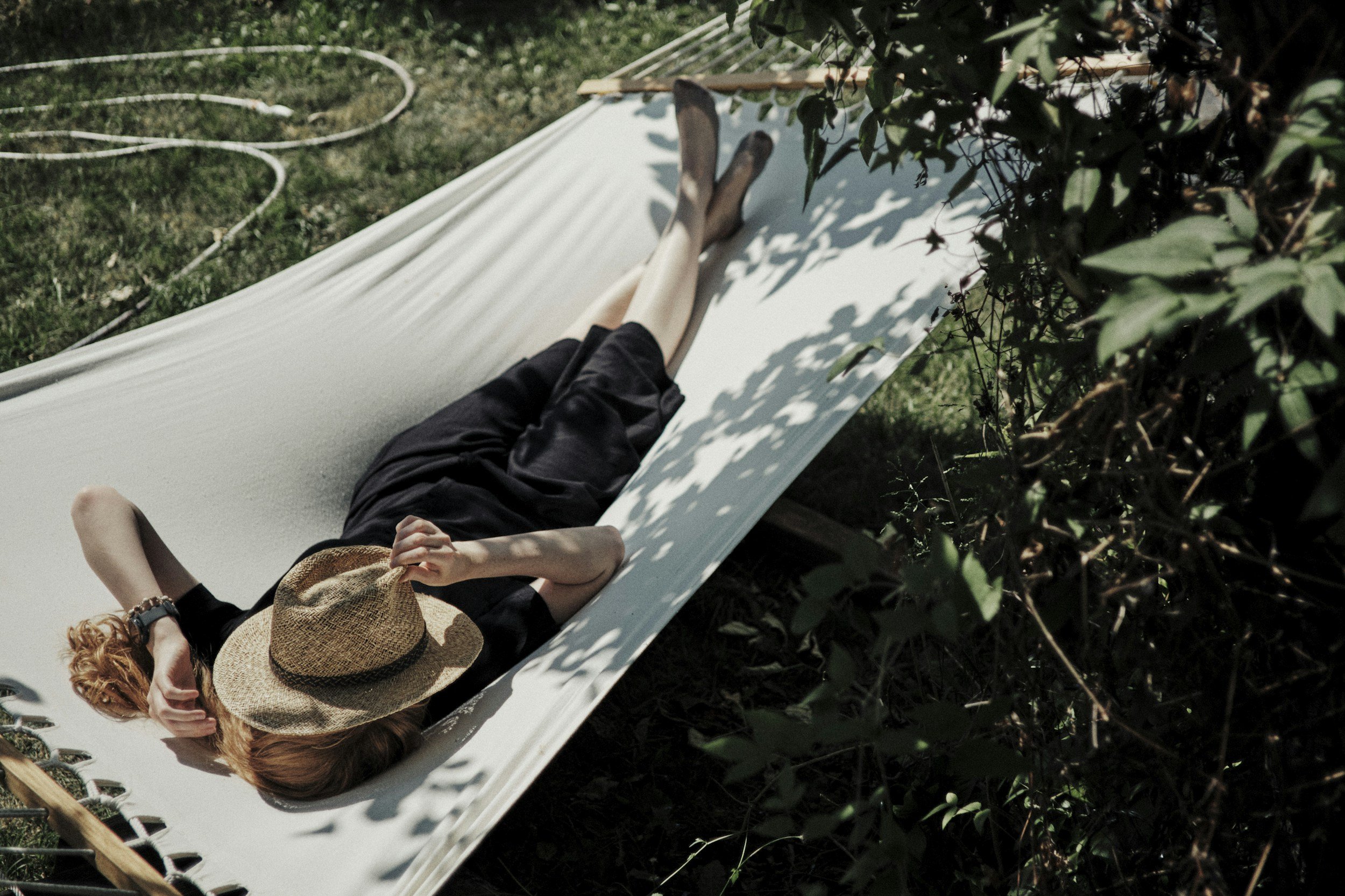 Person lying on a white bed or lounge outside, wearing a straw hat, black clothing, and a watch, with greenery and bushes around.