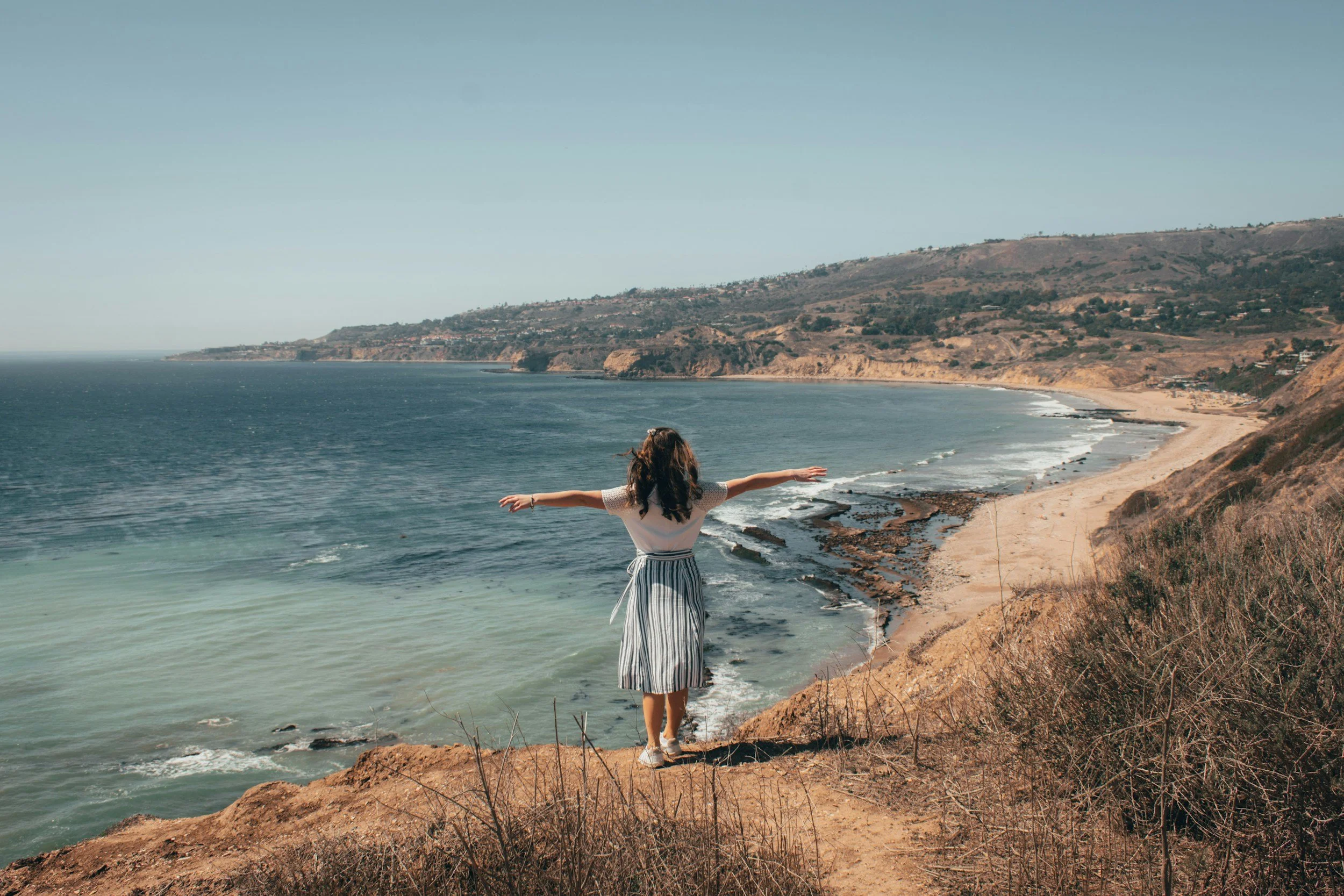 A woman stands on a cliff overlooking the ocean with her arms outstretched, facing the water and distant shoreline during daytime.