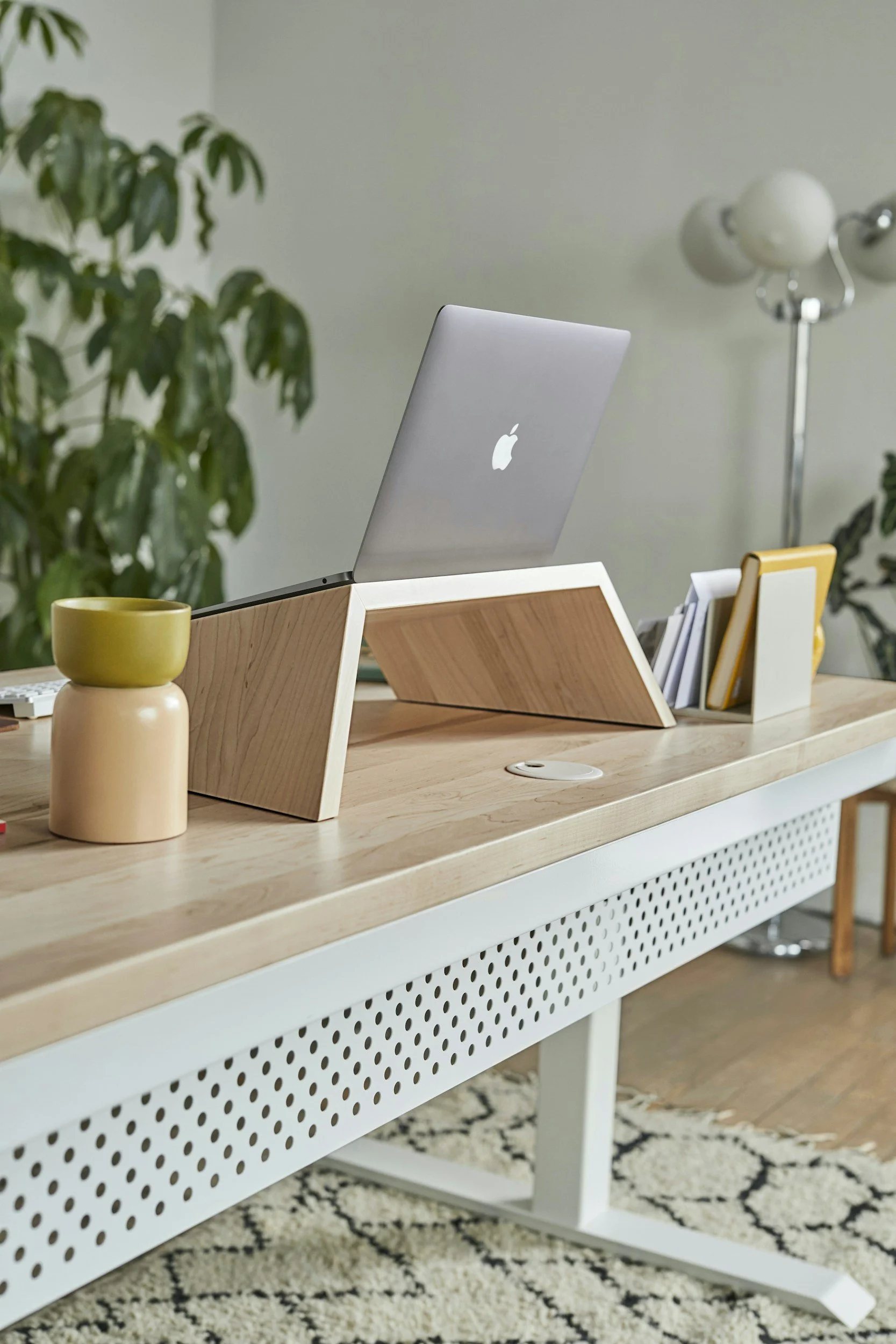 A modern office desk with a silver Apple MacBook on a wooden adjustable stand, a white mesh side panel, a yellow and white desk organizer with papers, a yellow ceramic cup, and a white wireless charging pad. In the background, there's a green potted plant, a tall floor lamp with multiple white and gray globes, and a patterned area rug.