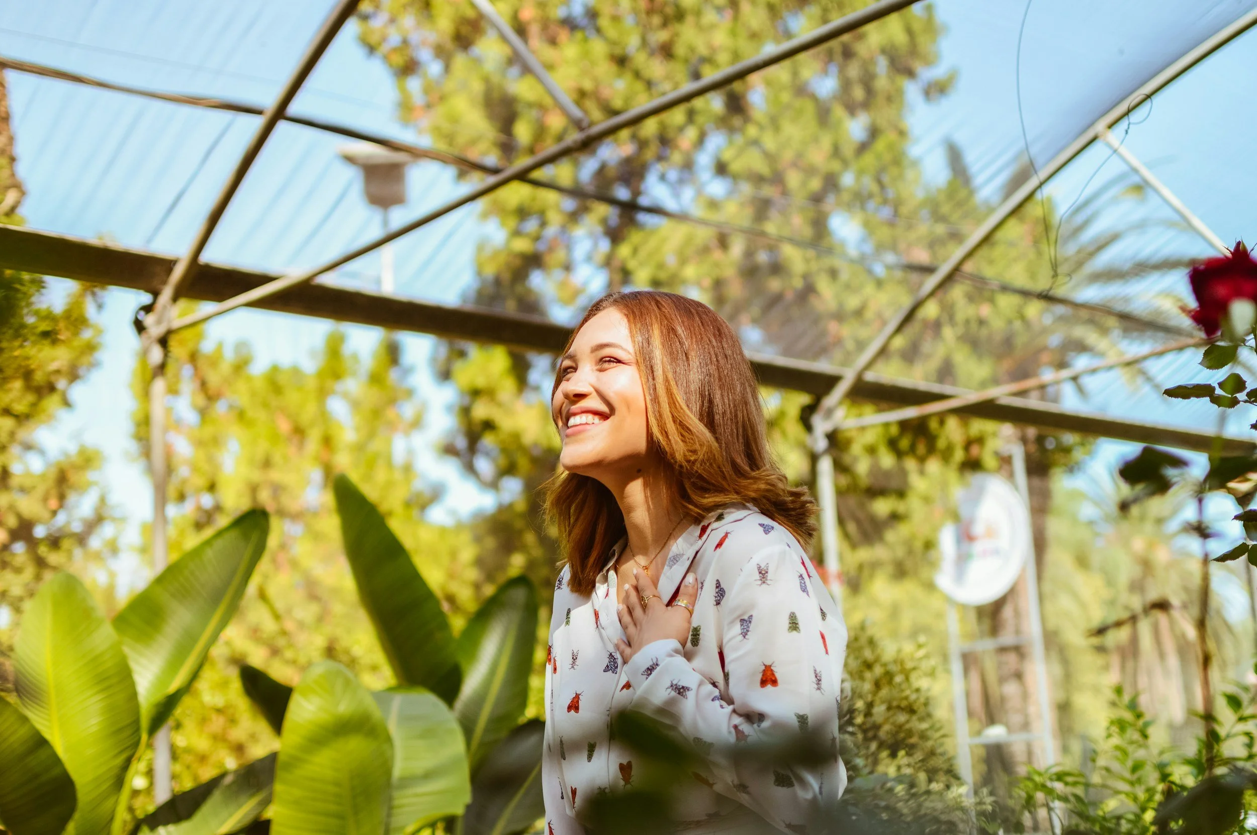 Woman smiling with hand on chest in greenhouse surrounded by lush green plants and trees, with a blue tarp overhead.
