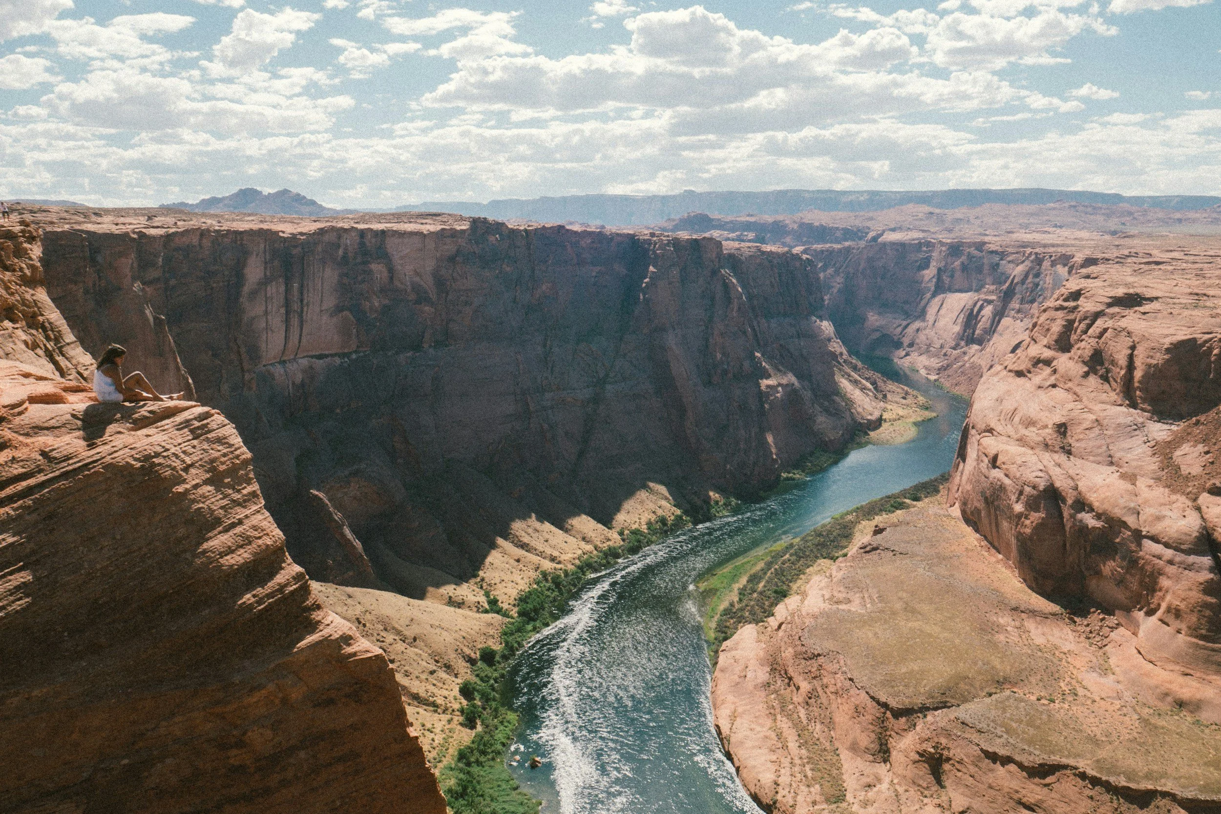 A woman sitting on a rocky ledge overlooking a canyon with a river flowing through it, under a partly cloudy sky.