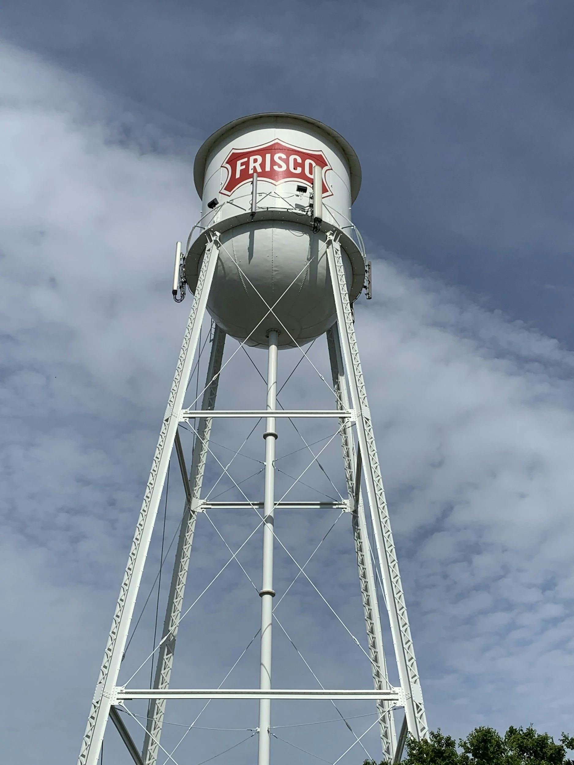 A tall white water tower with the red and white Frisco logo on it, set against a cloudy sky and some trees at the bottom.
