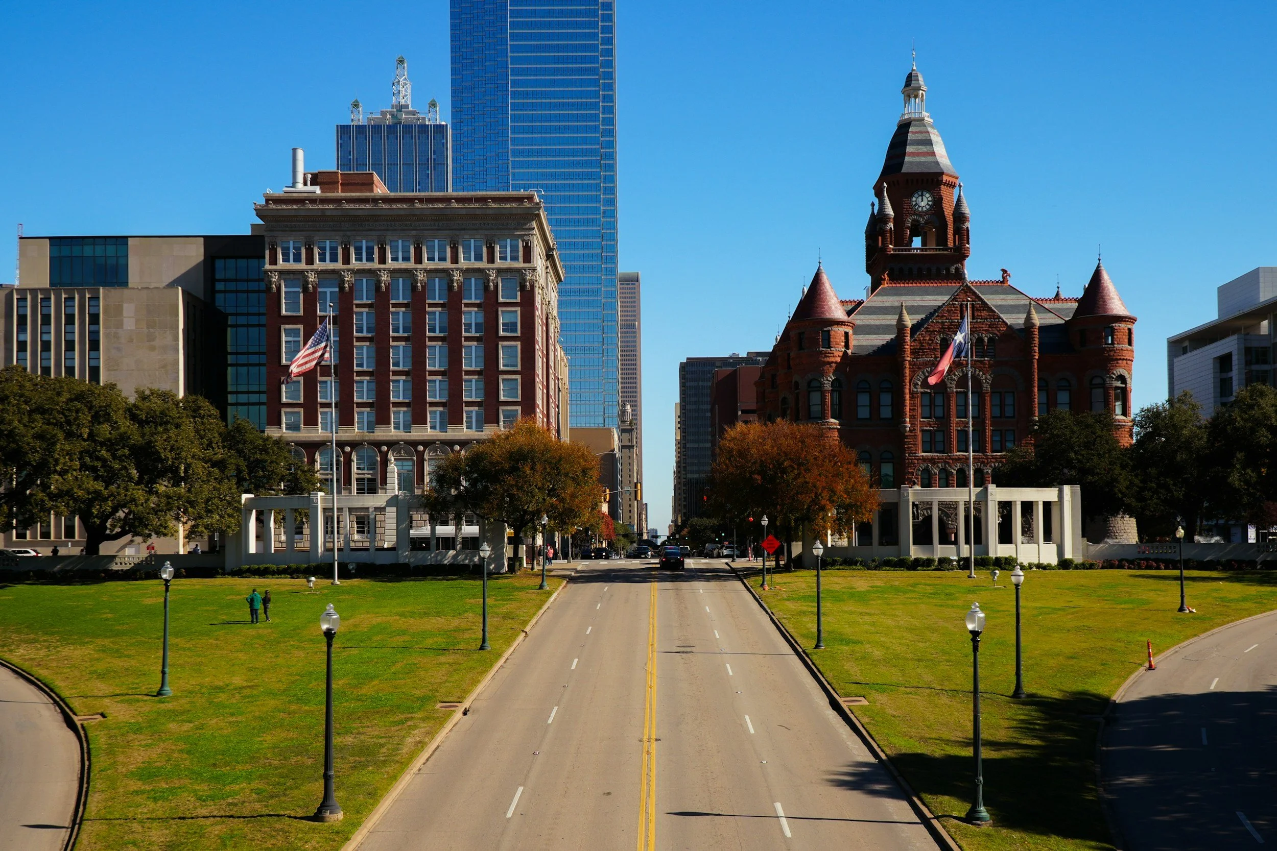 A city street with a park in the foreground, lined with trees and lamp posts. In the background, there are tall modern skyscrapers and a historic brick building with a clock tower under a clear blue sky.