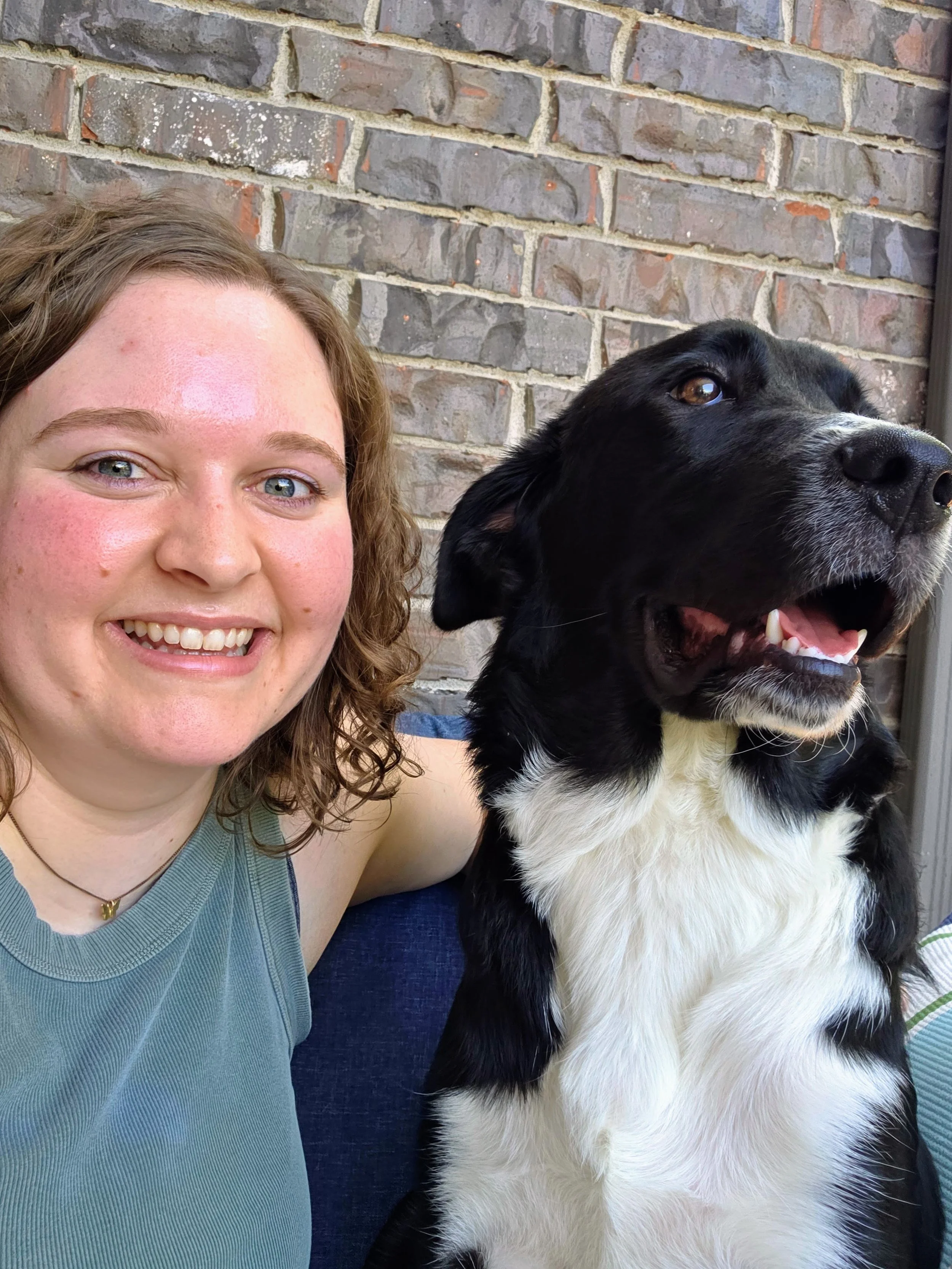 A woman with curly blonde hair, wearing a gray sleeveless top and a necklace, taking a selfie with a large black and white dog with its mouth open in front of a brick wall. The woman is smiling, and the dog appears happy.