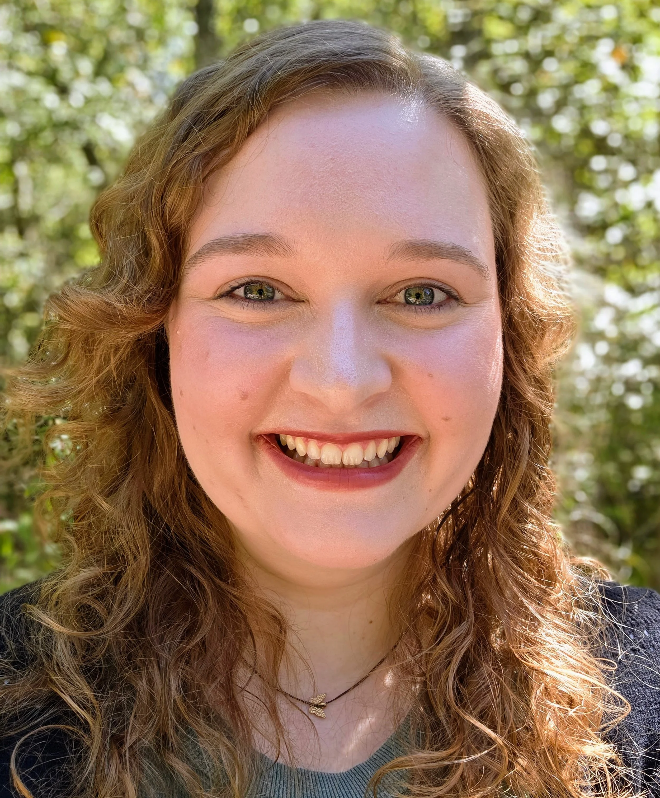 Close-up of a smiling woman's face outdoors with curly red hair and green eyes, wearing a necklace and a dark top, with blurred greenery in the background.