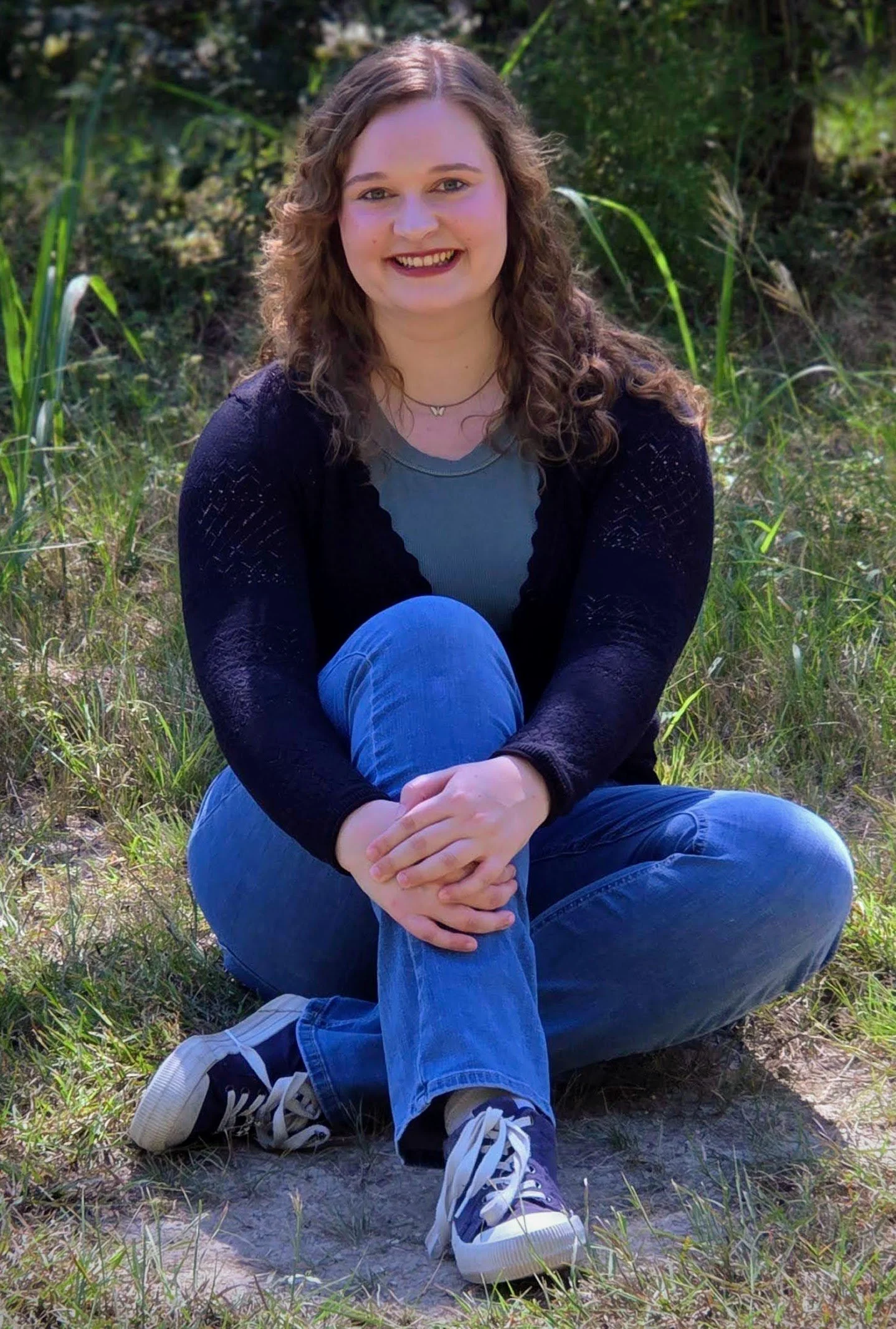 A young woman with curly hair sitting cross-legged outdoors in a grassy area, smiling at the camera, dressed casually in blue jeans, a gray shirt, a black cardigan, and sneakers.