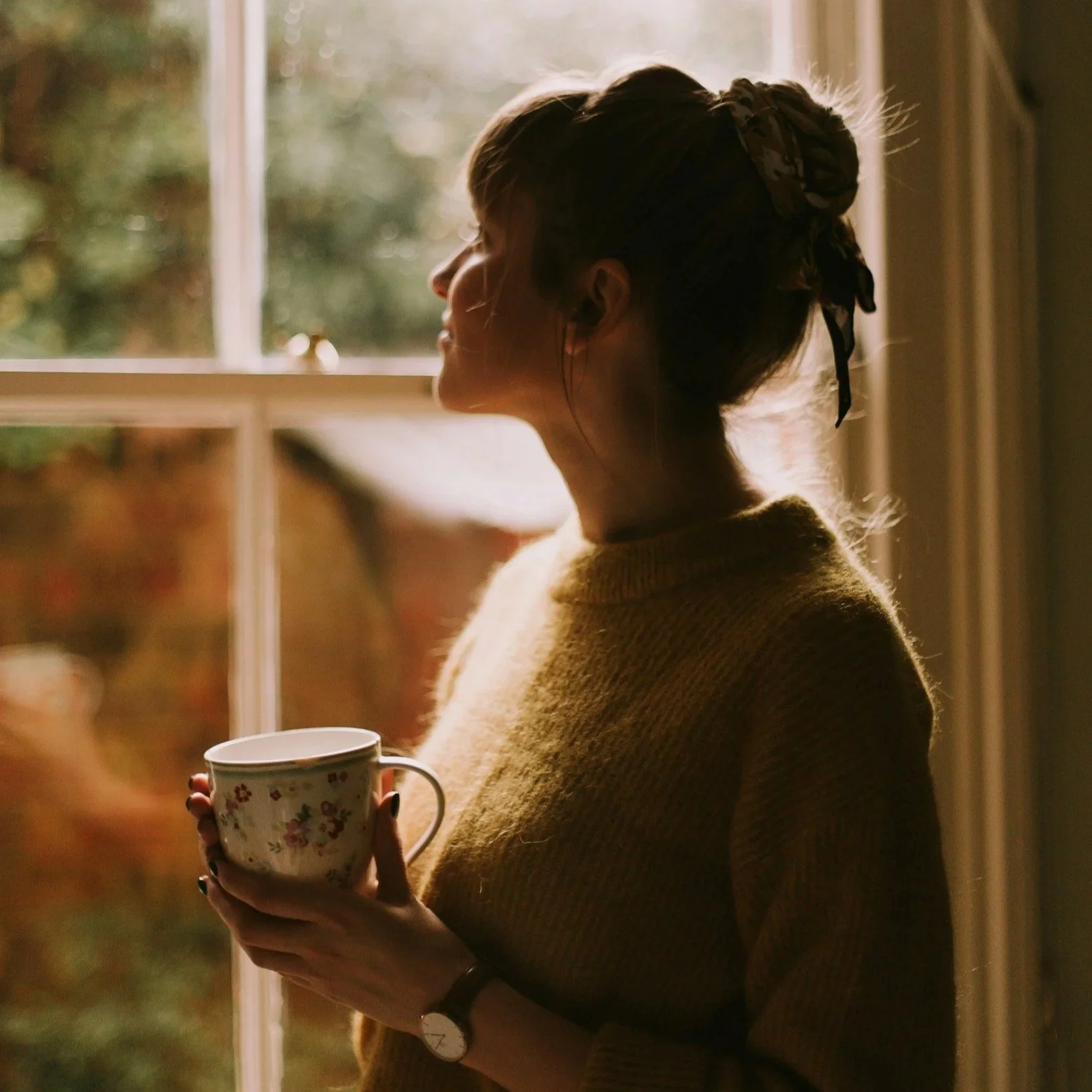 Woman in a yellow sweater staring t a sunny window while holding a floral teacup.
