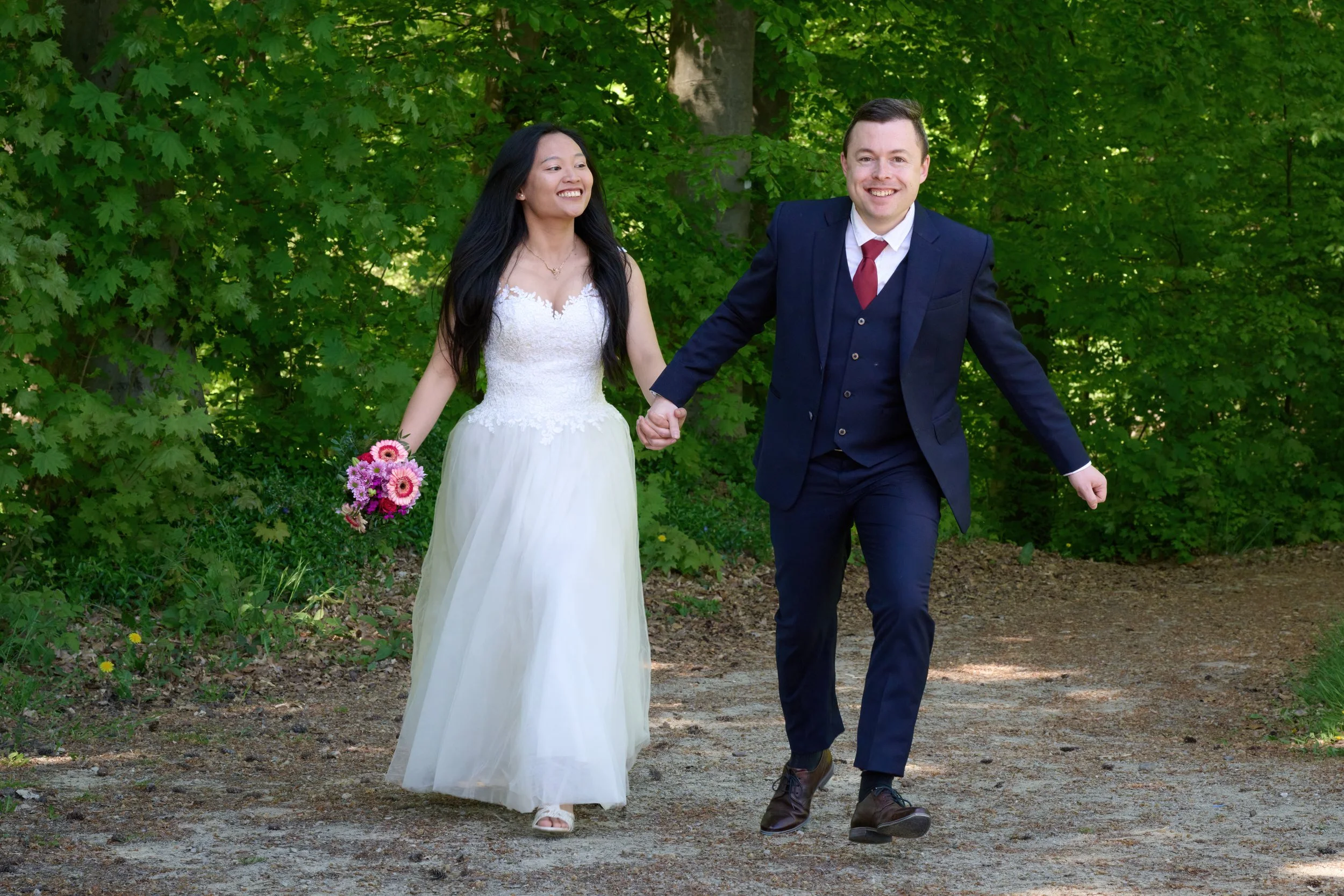 Ein glückliches Paar bei einer Hochzeit im Freien, das Hand in Hand durch einen Park mit grünen Bäumen geht.