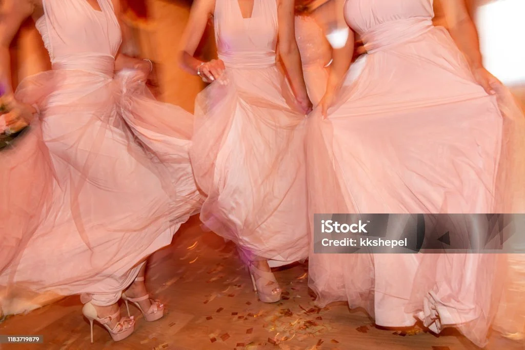 Damen in langen, rosa Kleidern tanzen auf einer Party oder Hochzeit, wobei ihre Kleider schwingen und glitzernde Konfetti auf dem Boden liegen.