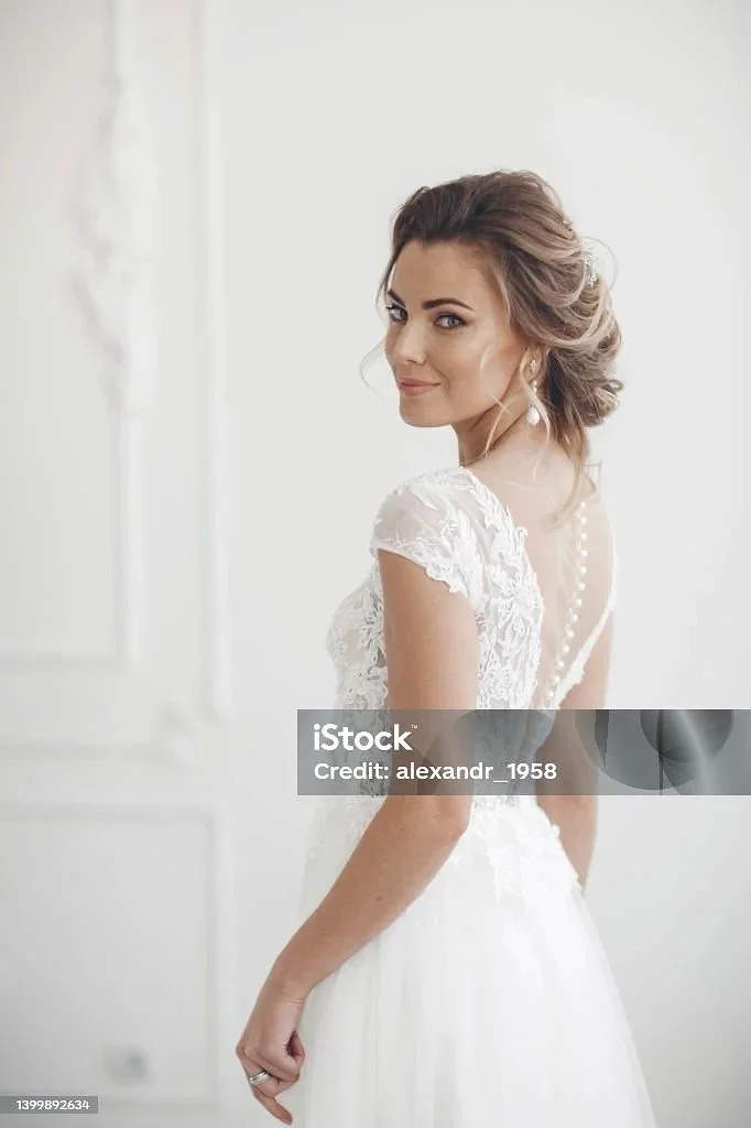 Eine Frau in einem weißen Brautkleid, die zum Rücken hin gedreht ist und in die Kamera lächelt.
