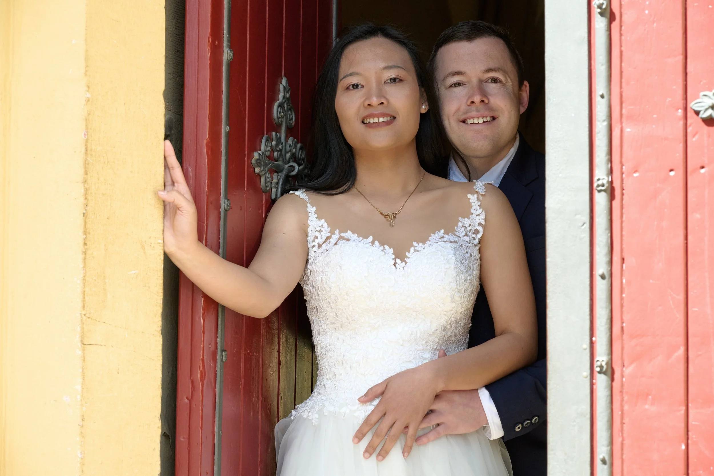 Ein Paar in Hochzeitskleidung schaut glücklich in die Kamera, stehend in einem Fensterrahmen.