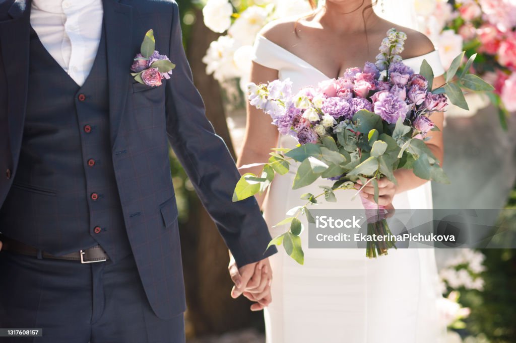 Ein Brautpaar hält Händchen während einer Hochzeit, die Braut trägt ein weißes Kleid und hält einen großen Blumenstrauß, der Bräutigam trägt einen dunklen Anzug mit Blumen im Revers.