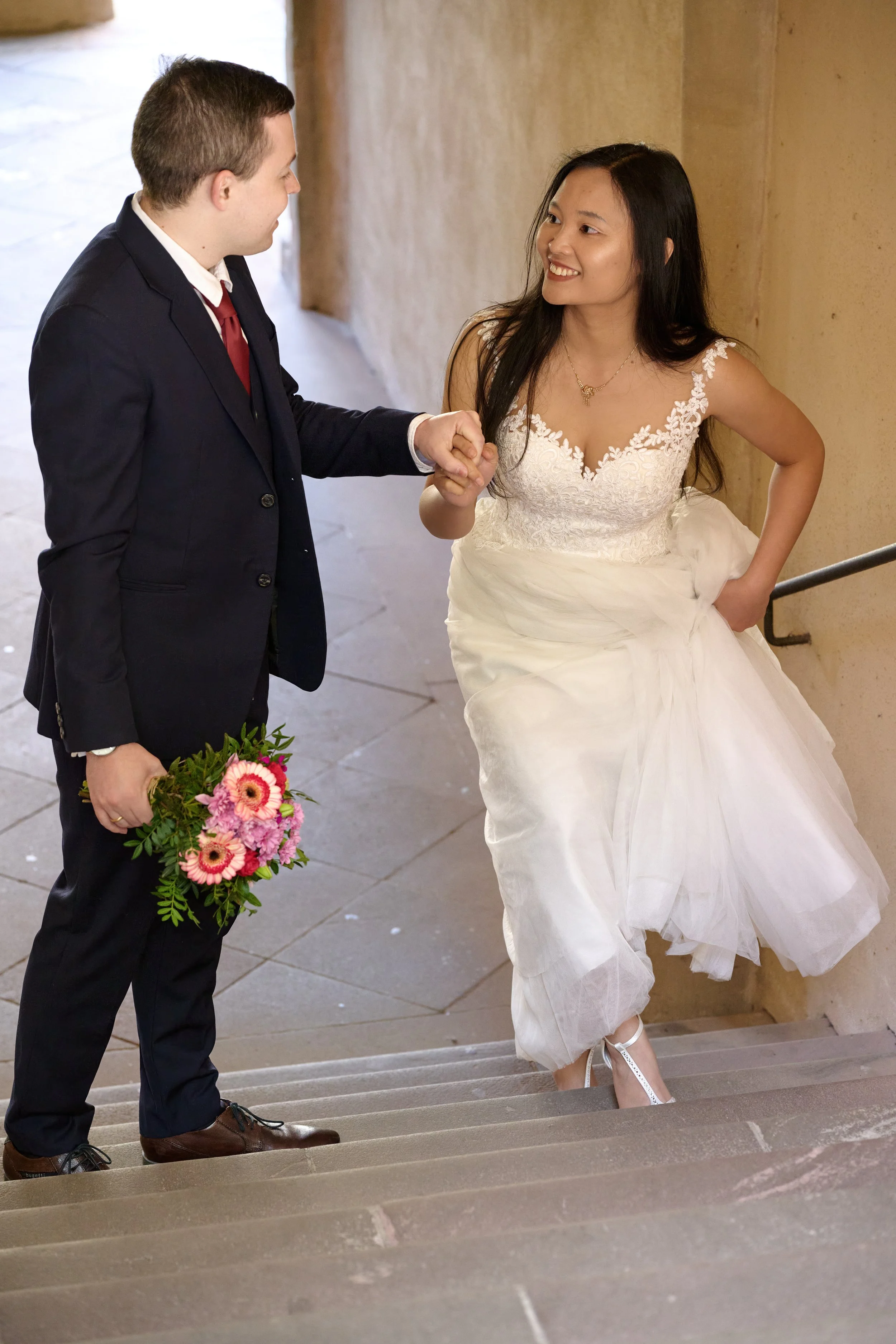 Ein Mann in Anzug hält die Hand einer Frau in einem weißen Brautkleid, während sie auf einer Treppe auf- oder absteigen. Er hält einen Blumenstrauß.