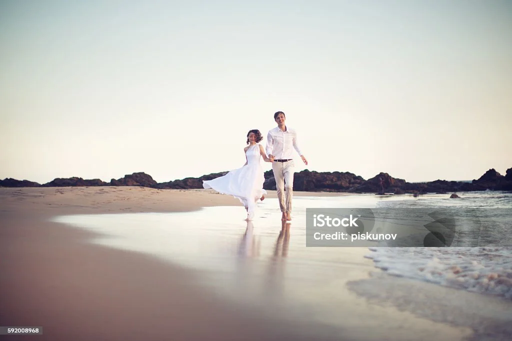 Ein Paar läuft Hand in Hand am Strand, die Frau trägt ein weißes Kleid, der Mann einen weißen Hemd und Hosen, im Hintergrund Felsen und das Meer.
