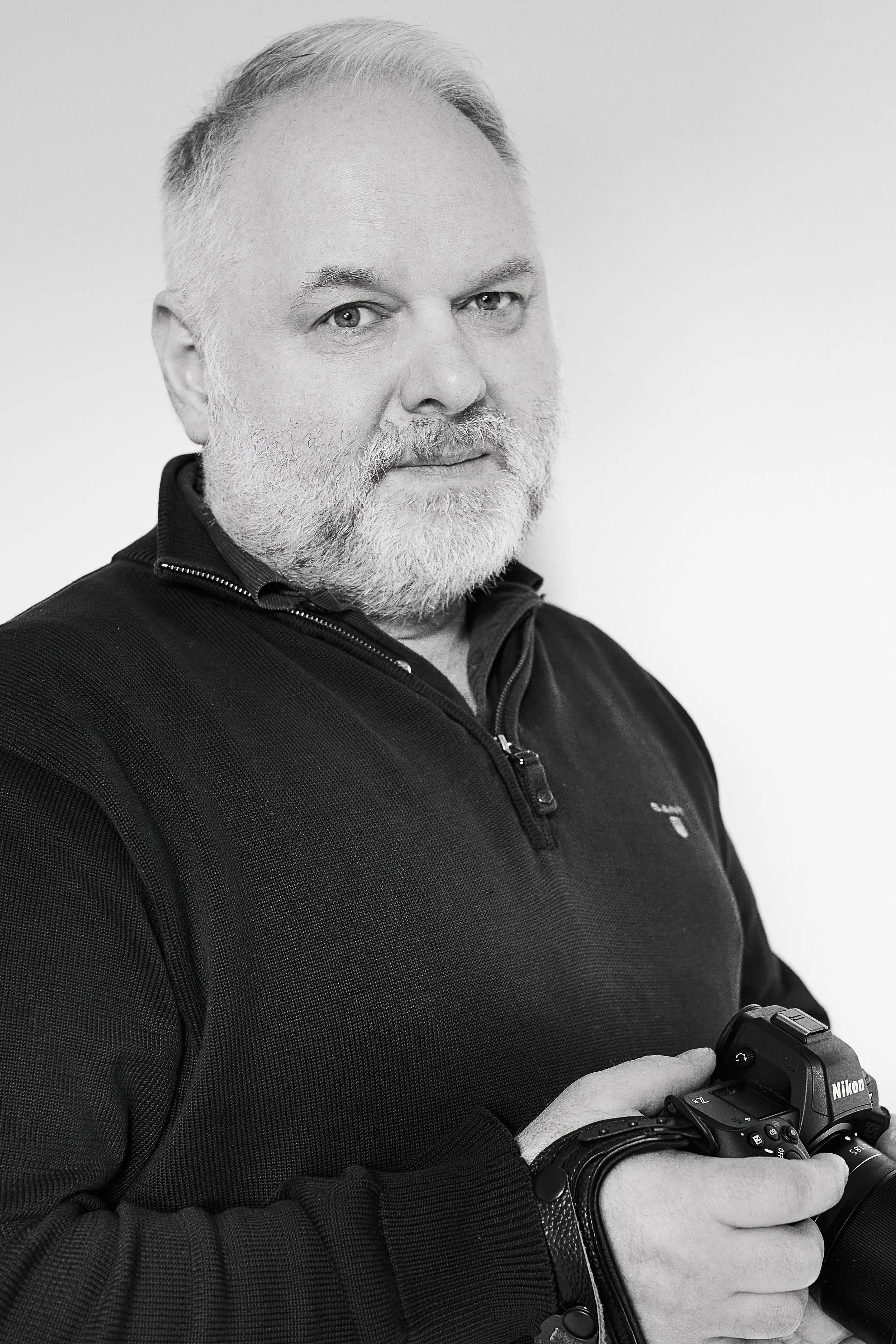 Mann mit grauem Bart und Haaren, trägt eine schwarze Jacke, hält eine Nikon Kamera in den Händen und scheint die Kamera zu inspizieren.
