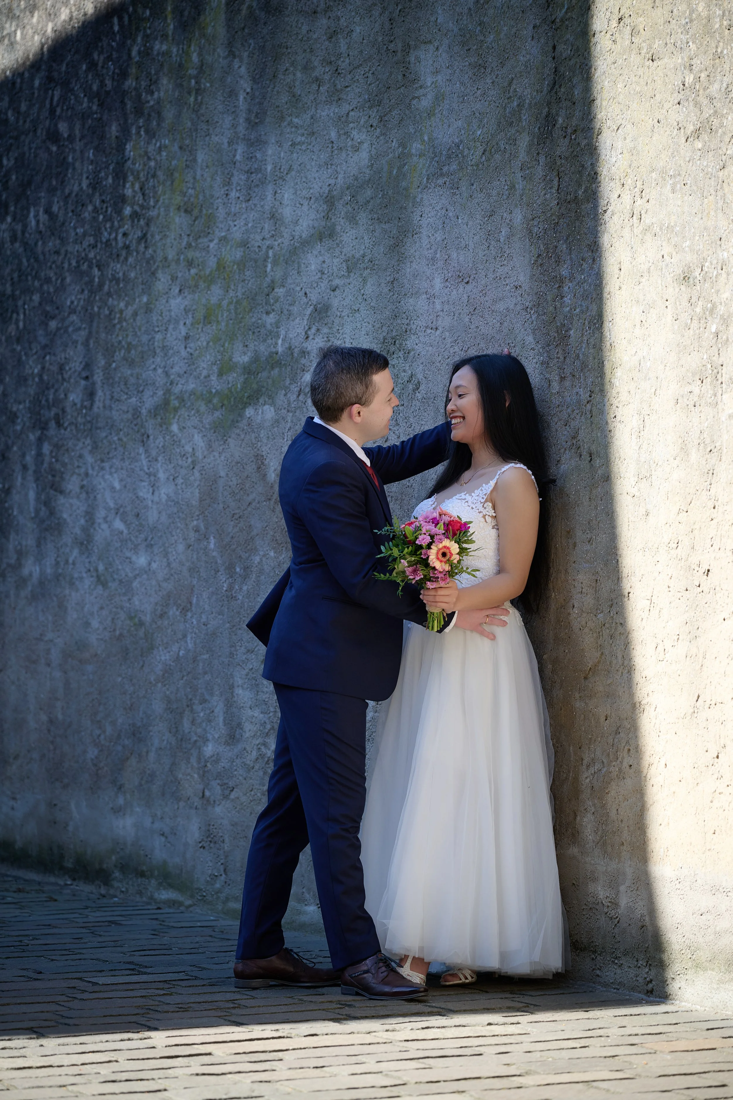 Ein Paar bei ihrer Hochzeit, steht vor einer grauen Steinwand, lacht und hält sich an den Händen, die Braut trägt ein weißes Brautkleid und hält einen Blumenstrauß, der Bräutigam trägt einen dunklen Anzug.