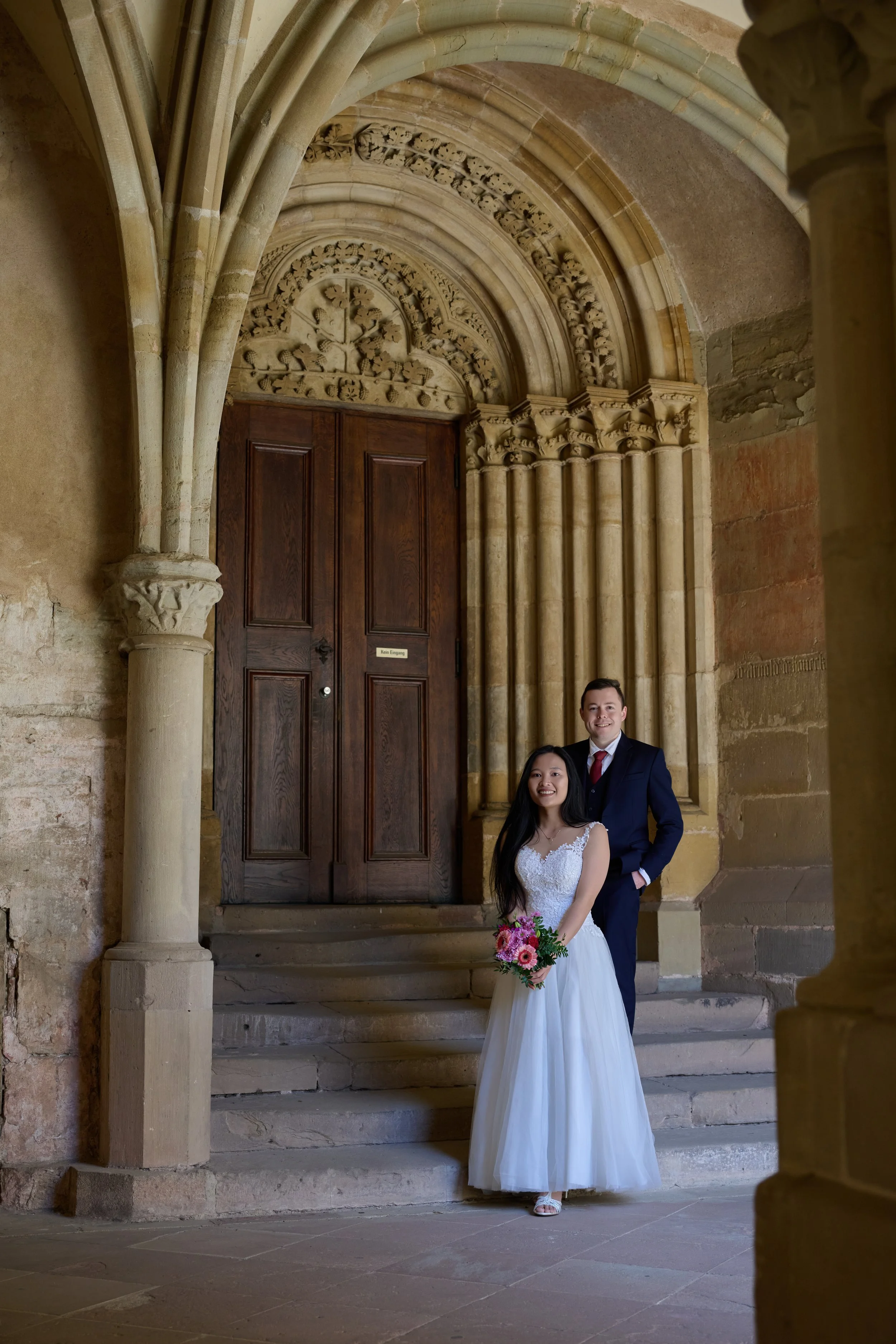 Braut in weißem Kleid und Blumenstrauß, Bräutigam in Anzug, stehen vor einer alten Steintür in einer historischen Kirche oder Burg, umgeben von steinernen Säulen und Bögen.