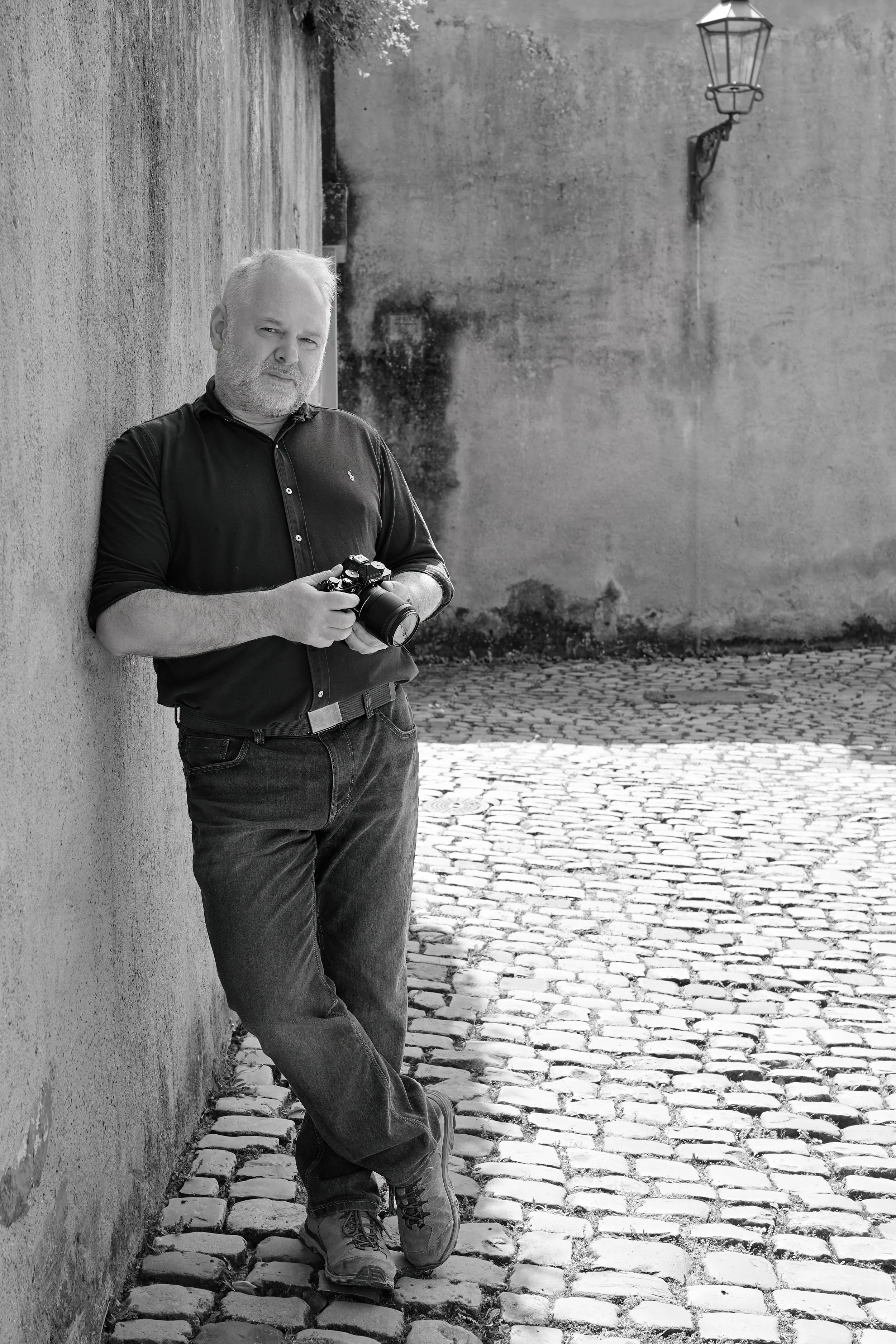 Mann mit weißen Haaren und Bart, in dunklem Hemd, lehnt an einer Wand mit einem Kamera in der Hand auf einer Kopfsteinpflasterstraße in Schwarz-Weiß-Fotografie.