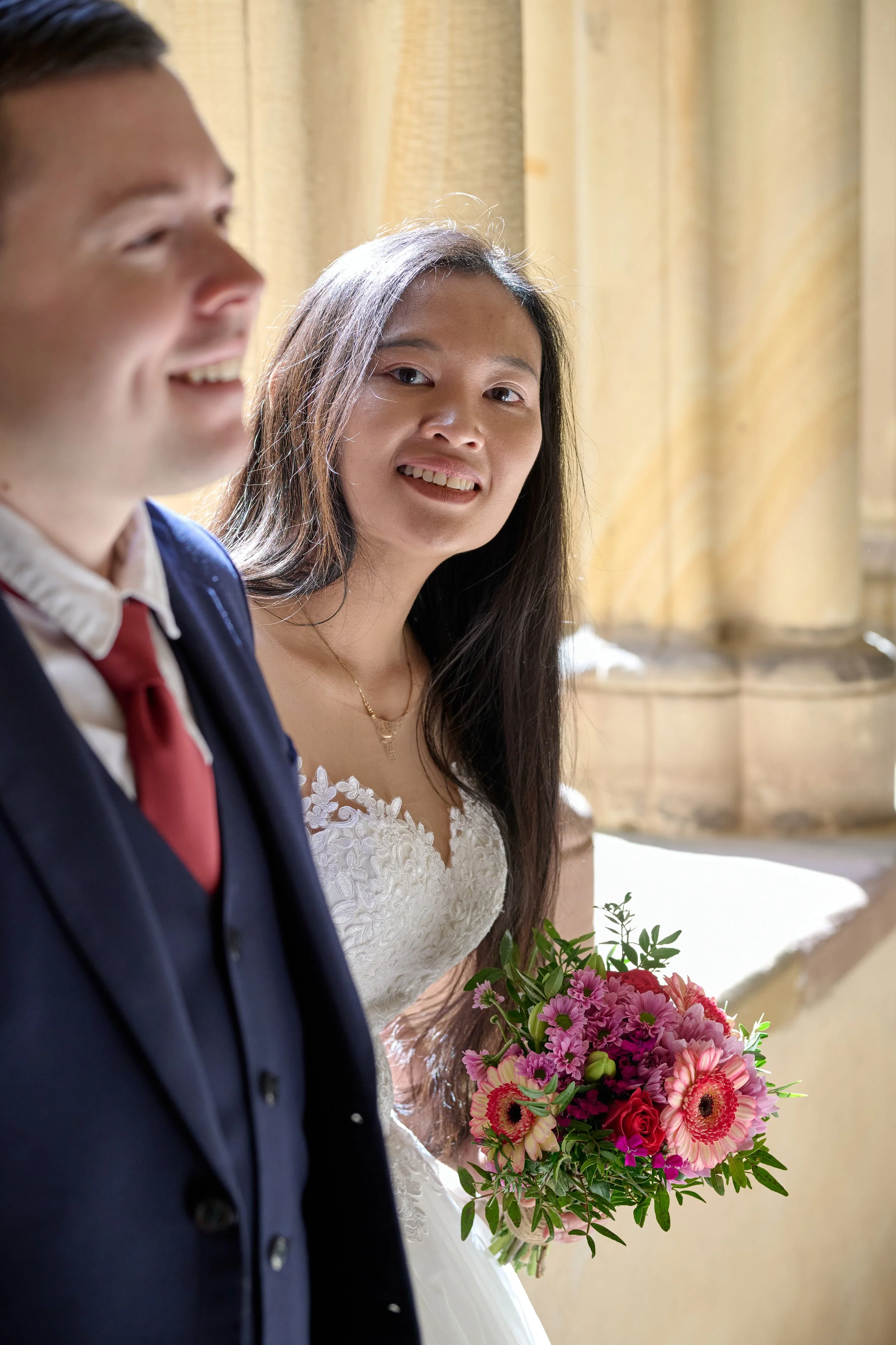 Eine Braut in einem weißen Hochzeitskleid hält einen Bouquet aus pinken und roten Blumen, während eine Person im dunklen Anzug neben ihr steht.