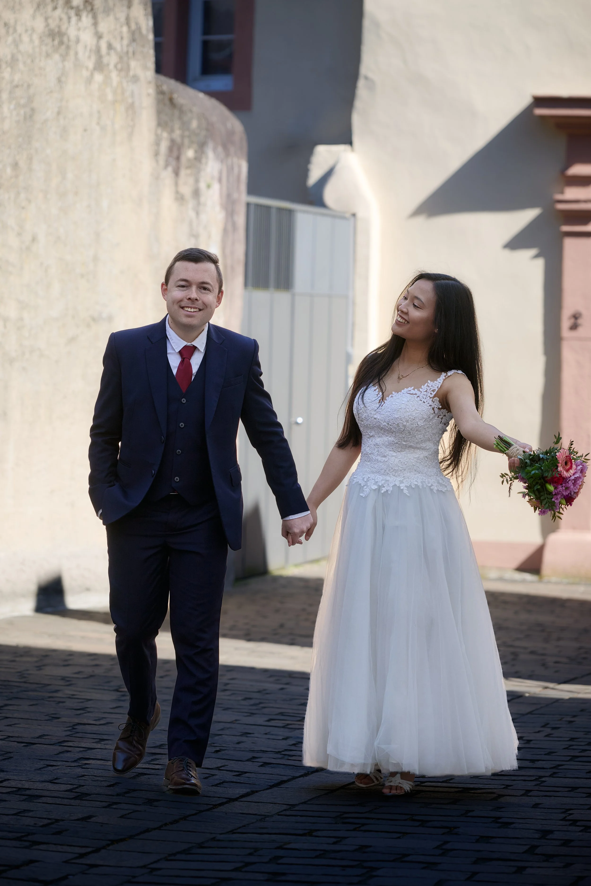 Ein Mann und eine Frau, die bei einer Hochzeit Händchen halten und vor einer Kulisse mit alten Gebäuden stehen.