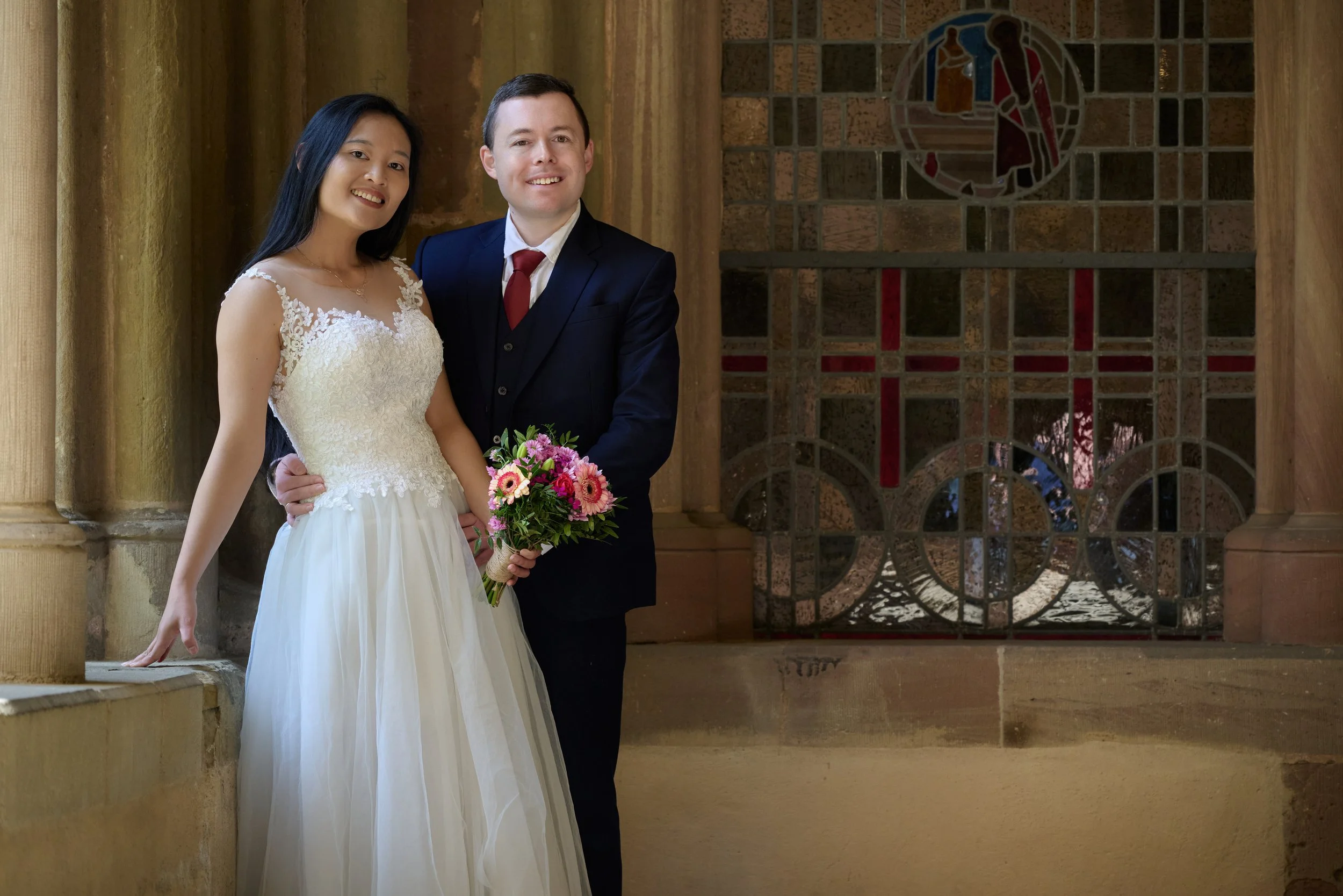 Ein Paar bei ihrer Hochzeit, das in einer Kirche steht. Die Braut trägt ein weißes Hochzeitskleid mit Spitze, der Bräutigam einen dunklen Anzug mit roter Krawatte. Die Braut hält einen Blumenstrauß, beide lächeln und sehen in die Kamera. Hinter ihnen