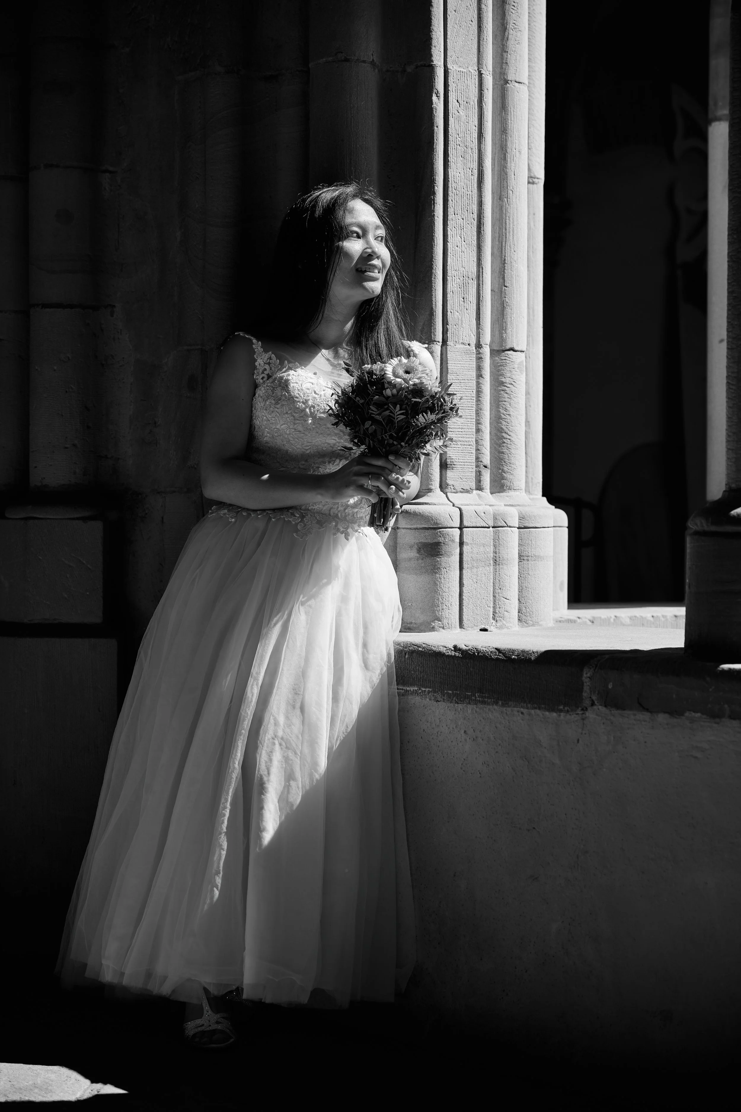 Eine lachende Frau in einem Brautkleid hält einen Blumenstrauß und steht neben einer Säule auf einer stone Bauwerk. Schwarz-weiß Fotografie, dramatisches Licht.