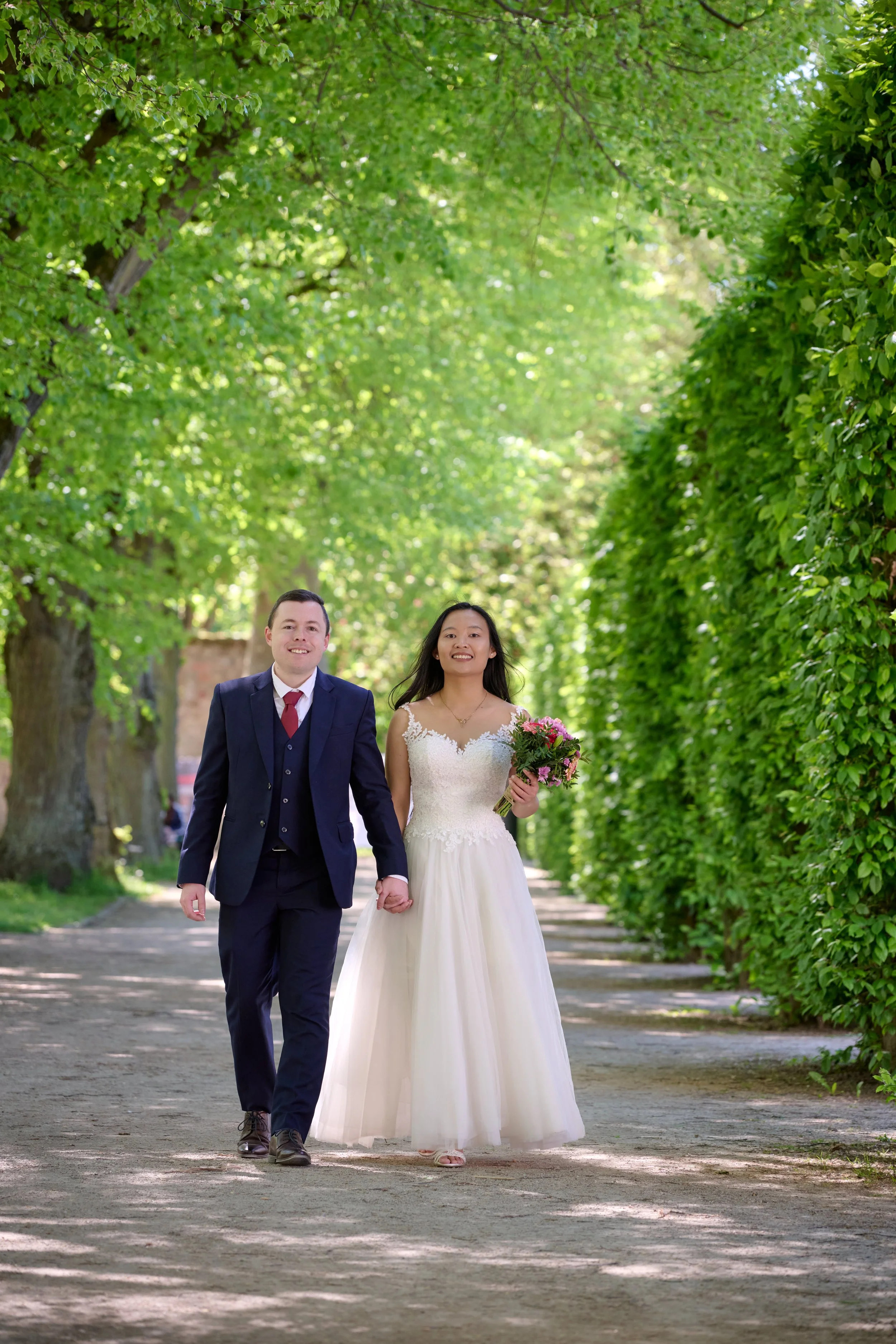 Ein Brautpaar, das Hand in Hand in einem Park mit grünen Bäumen spaziert, die Braut trägt ein weißes Kleid und hält einen Blumenstrauß, der Bräutigam trägt einen dunklen Anzug.