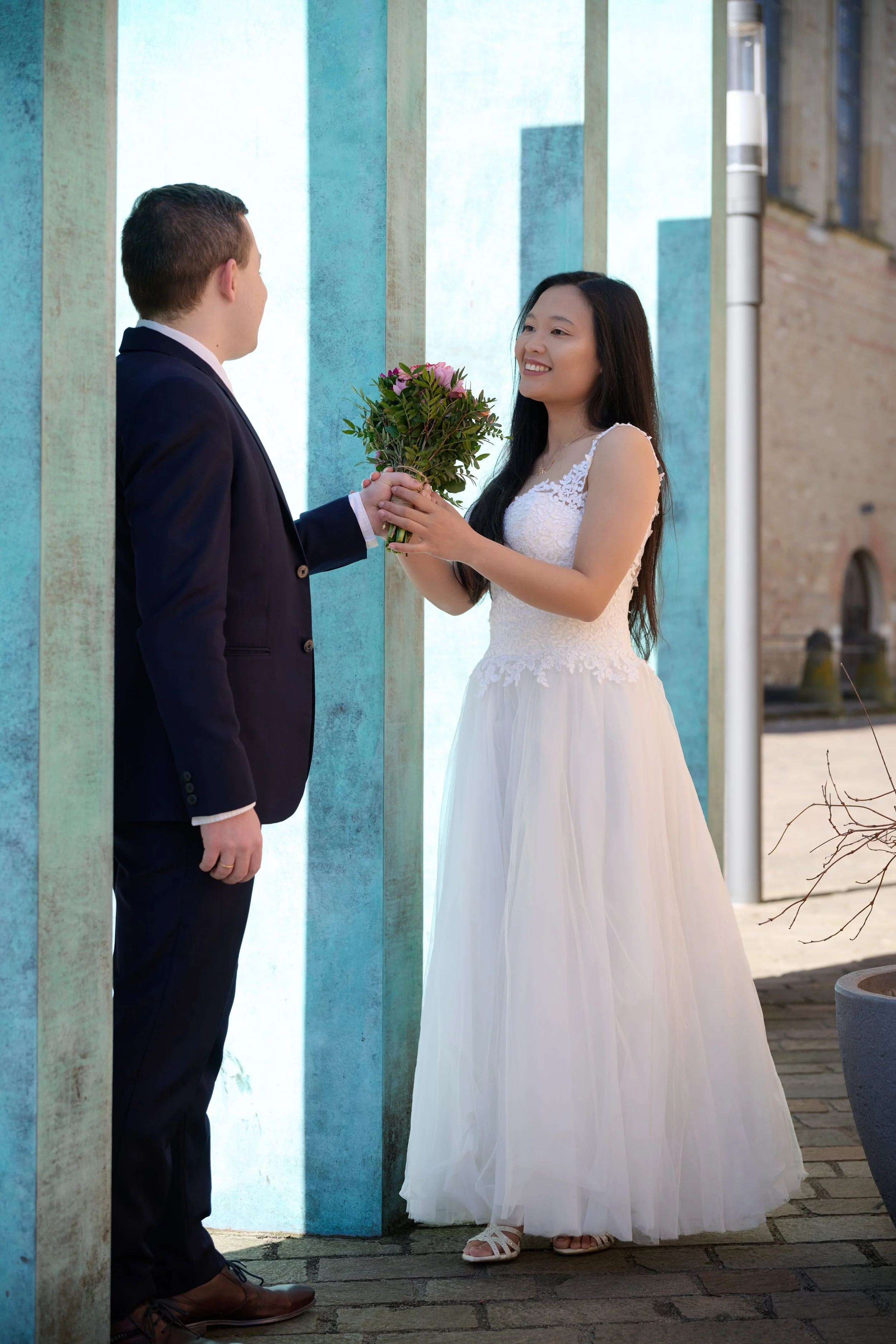 Ein Mann in einem Anzug überreicht einer lächelnden Frau in einem weißen Brautkleid einen Blumenstrauß, während sie sich gegenüberstehen.