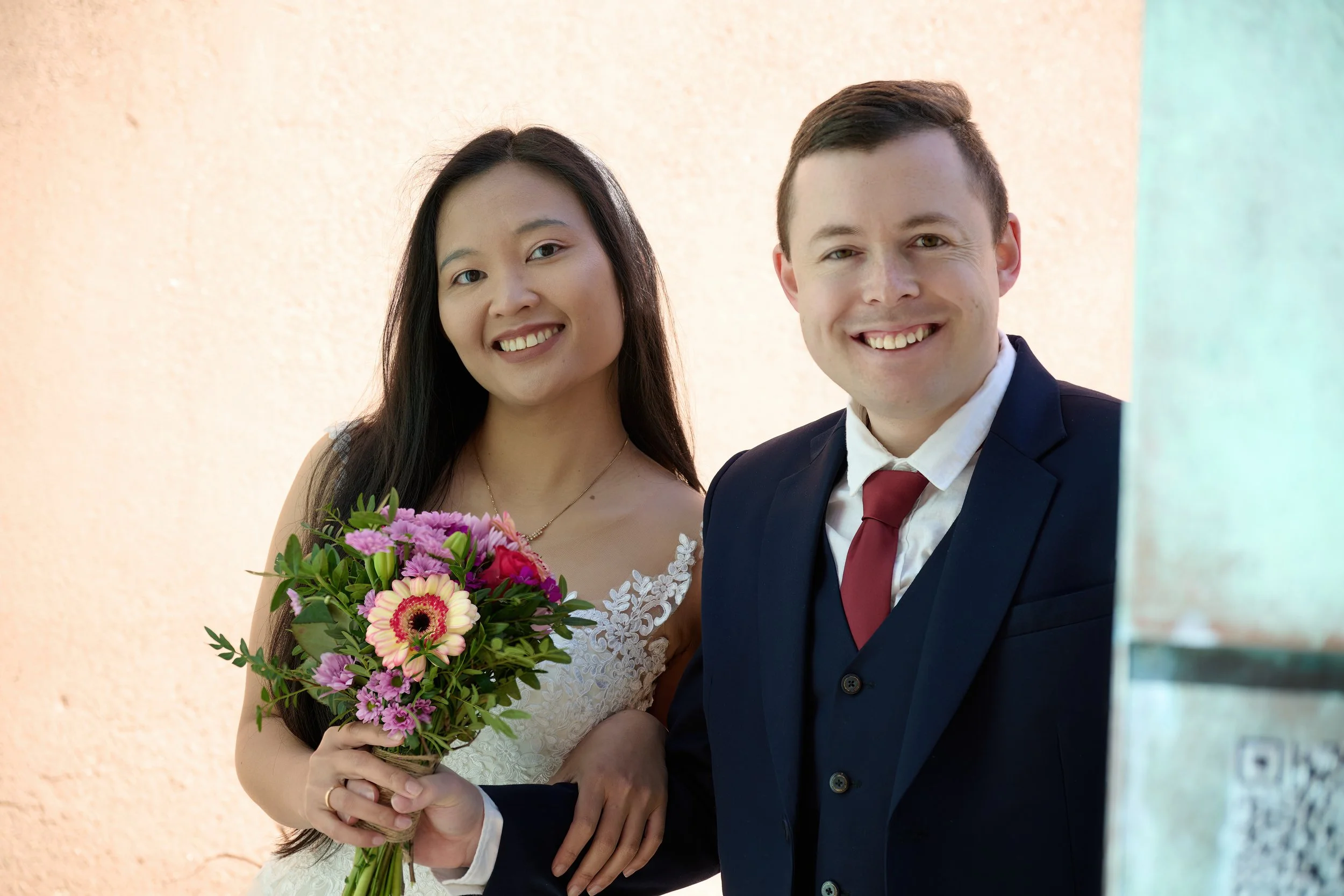 Ein glückliches Paar bei einer Hochzeit, die Braut hält einen Blumenstrauß, beide lächeln in die Kamera.