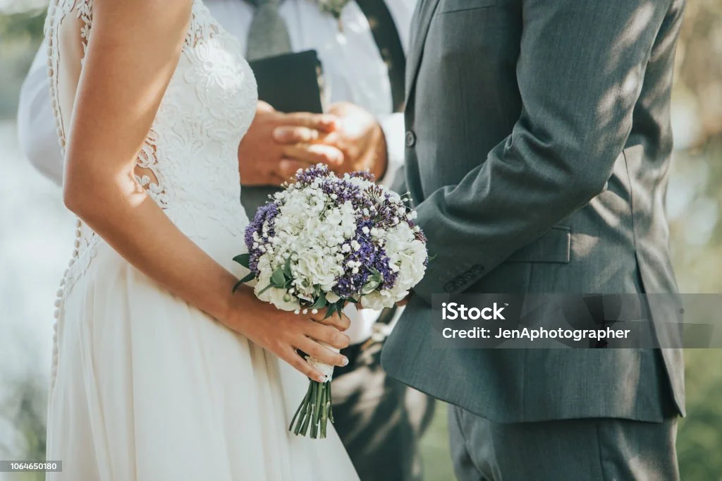 EinBraut und einBräutigam halten sich bei einer Trauung an den Händen, die Braut trägt ein weißes Hochzeitskleid und hält einen Blumenstrauß, der Bräutigam trägt einen grauen Anzug.