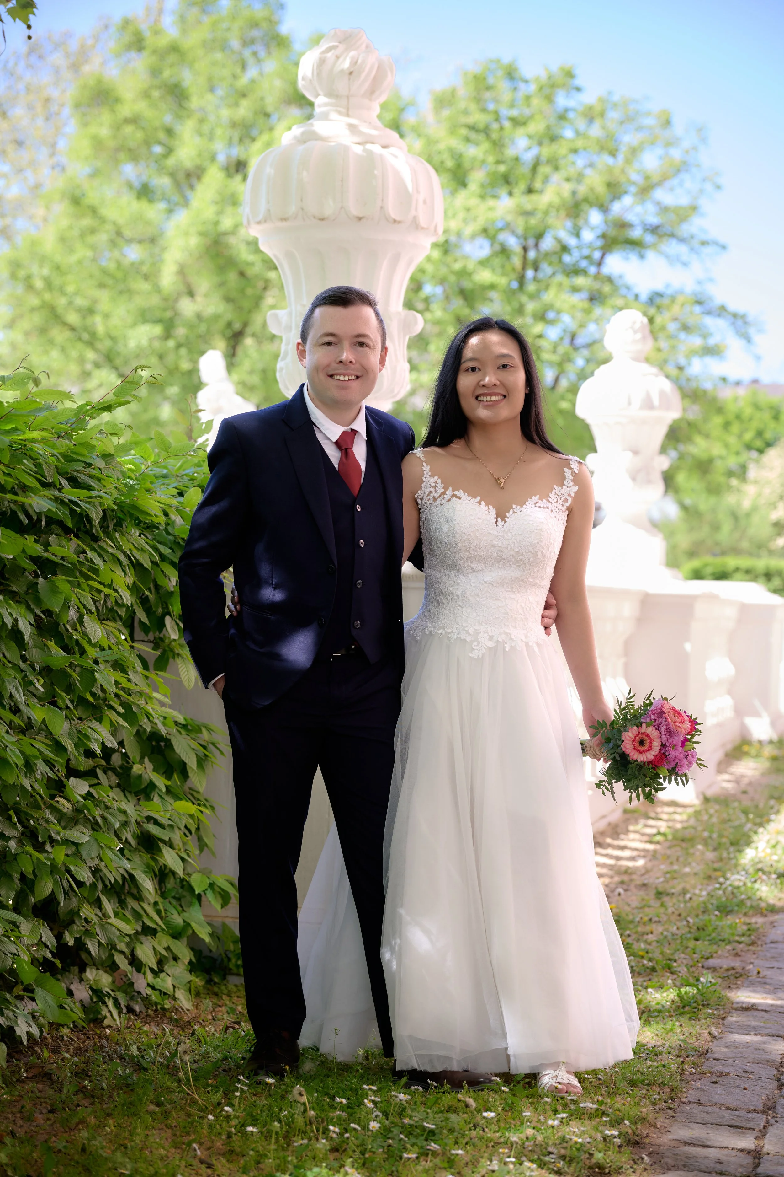 Ein Paar an einem sonnigen Tag im Freien, vermutlich anlässlich einer Hochzeit, stehend auf einem Gartenweg mit grünem Laub und blühenden Büschen im Hintergrund.