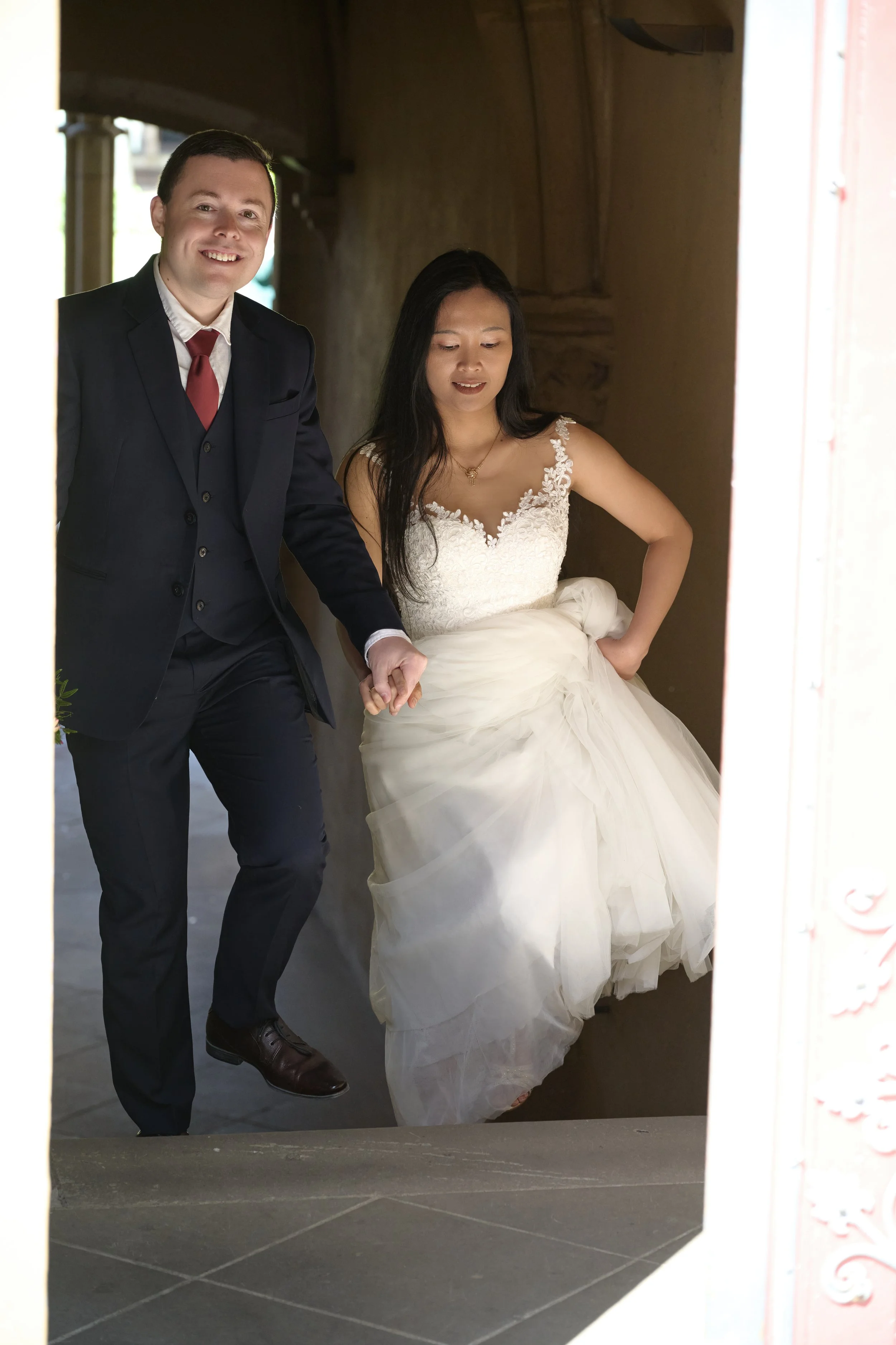 Ein Brautpaar, in einem dunklen Raum, die Braut trägt ein weißes Hochzeitskleid, der Bräutigam trägt einen dunklen Anzug mit roter Krawatte. Sie halten Händchen und posieren für einen Spiegel
