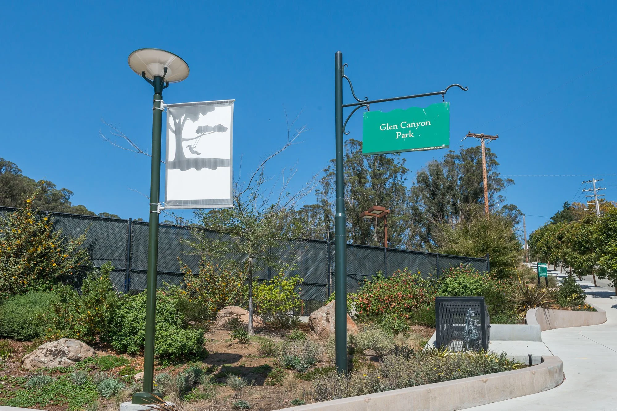 Park entrance sign for Glen Canyon Park with trees, bushes, rocks, and a sidewalk under a blue sky.