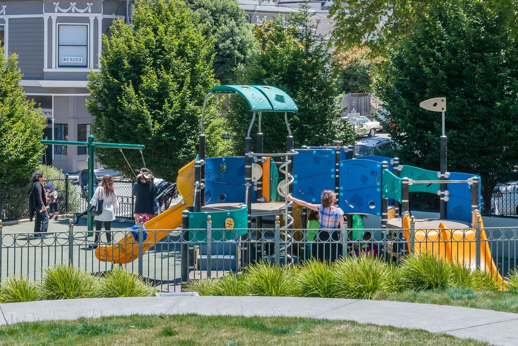 Children and adults at a colorful playground with slides and climbing structures, surrounded by trees and a fence.