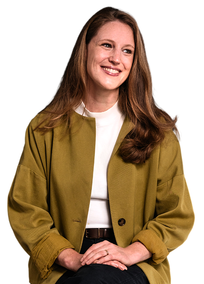 Ein lächelnder Frau mit braunen Haaren, die einen grünen Mantel und einen weißen Pullover trägt, sitzt vor schwarzem Hintergrund.