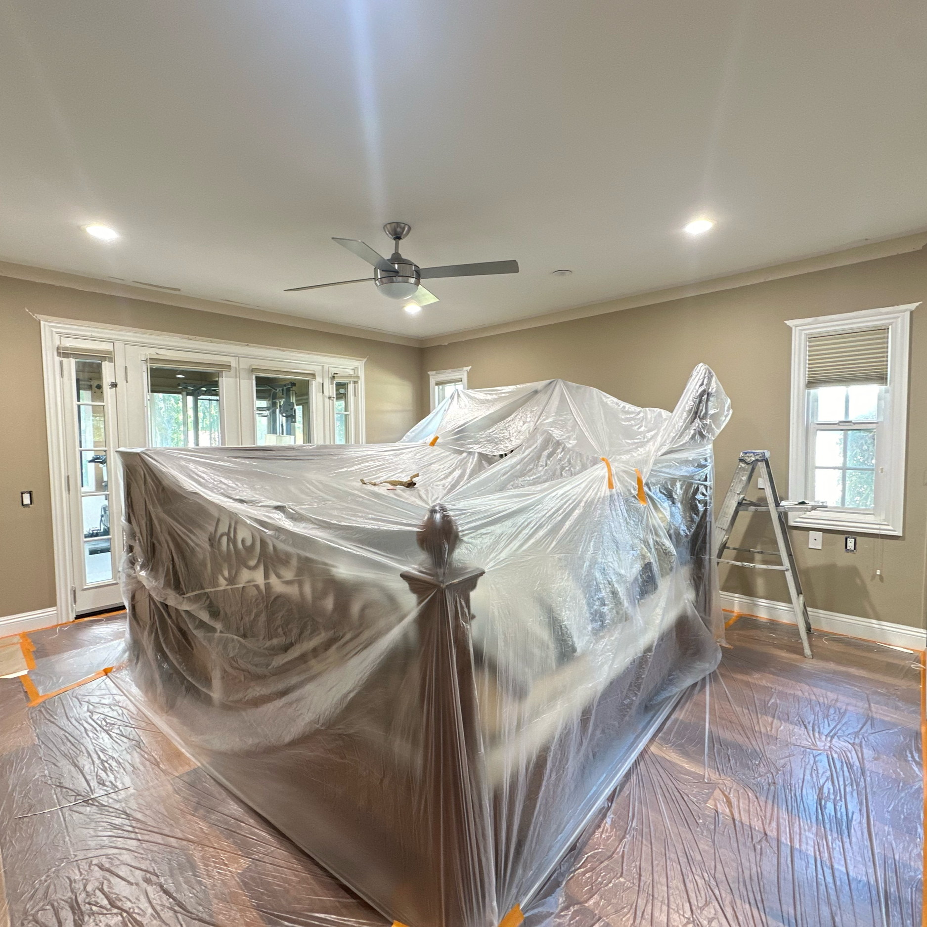 Living room covered with plastic sheeting and painter's tape, indicating ongoing painting or renovation work, with a ladder on the right and ceiling fan overhead.